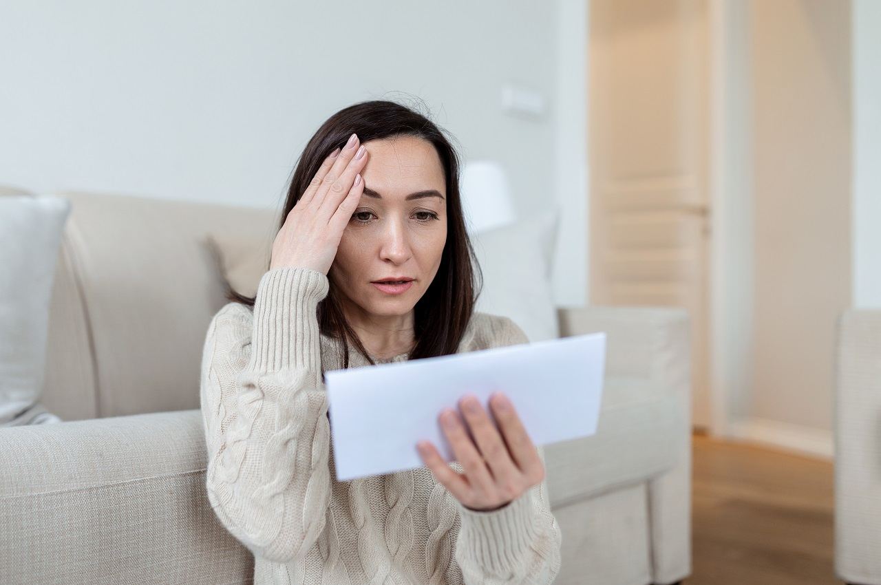 Mulher com expressão preocupada segurando carta de inadimplência financeira e apoiando a mão na cabeça. Ela veste suéter bege e está sentada no chão com as costas apoiadas no sofá.