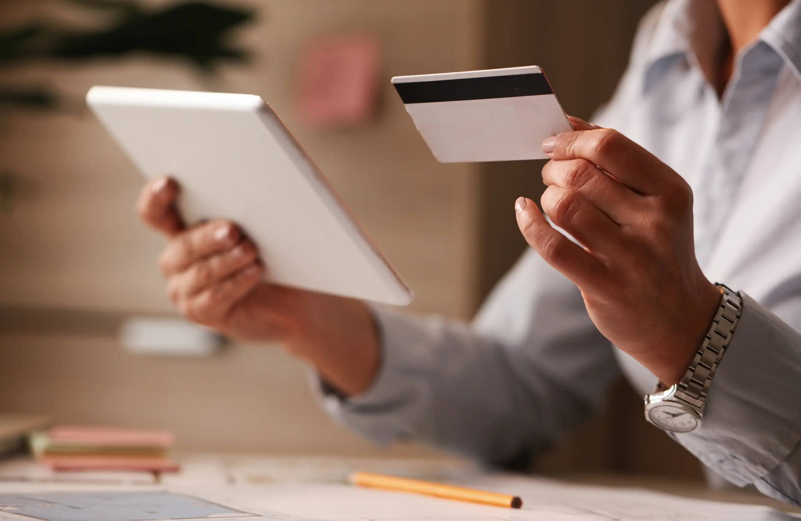 Mãos segurando um tablet e um cartão de crédito sobre uma mesa de escritório. O rosto da pessoa não está visível. 