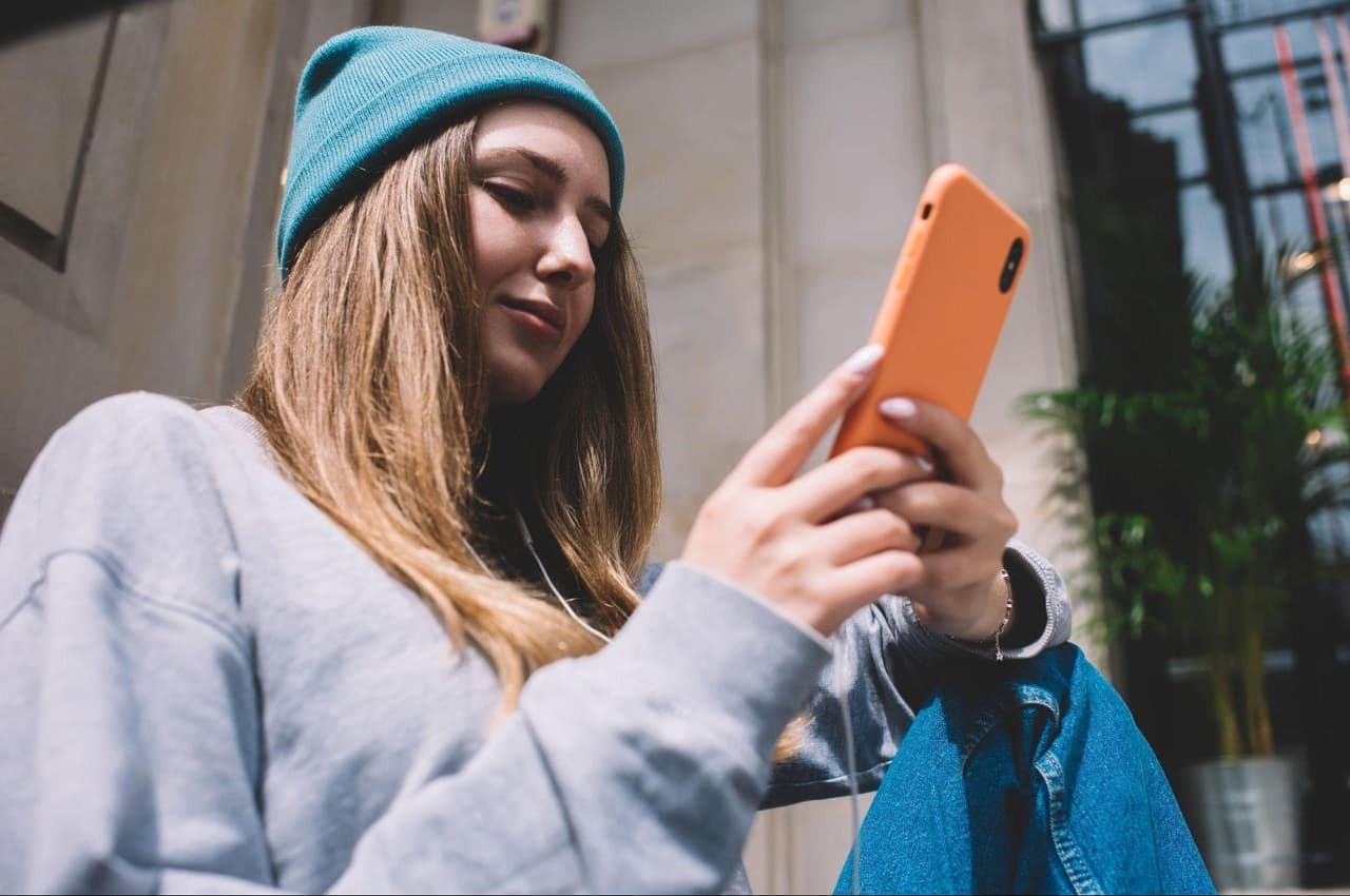 Mulher de cabelos loiros, touca de frio laranja e moletom cinza, acessando empréstimo FGTS pelo app BV no celular.