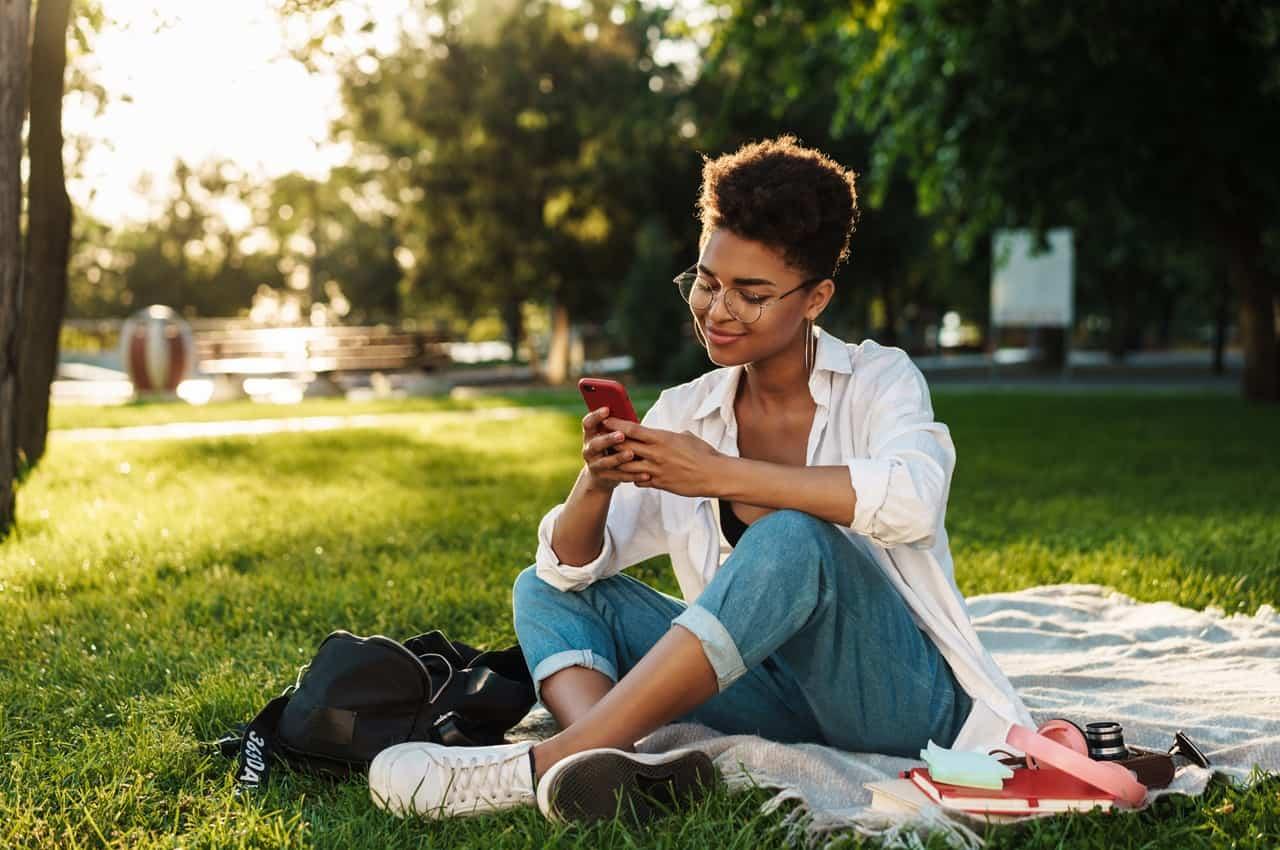 Mulher sorridente sentada no gramado de uma praça arborizada. Ela utiliza o celular, usa óculos de grau e veste calça jeans e cropped preto.