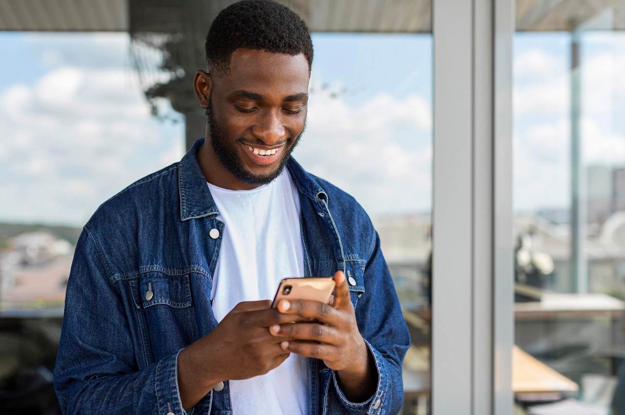 Homem sorridente, em pé, acessando FGTS Completão BV pelo celular. Ele veste camisa branca básica, jaqueta jeans e tem cabelos curtos pretos.