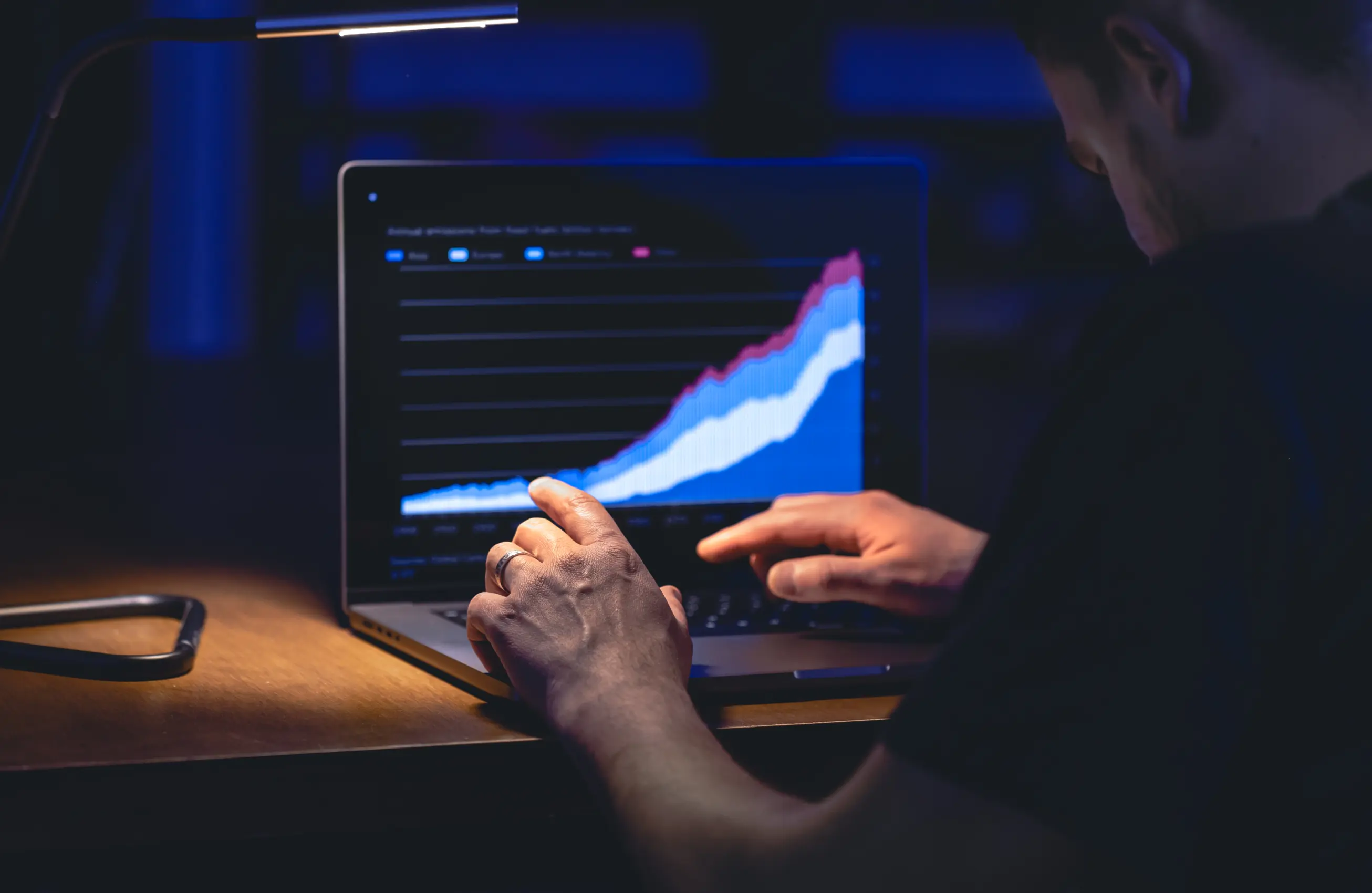 Pessoa olhando para um laptop em ambiente escuro, com gráfico em tela que mostra linhas ascendentes e áreas coloridas; as mãos da pessoa estão próximas ao teclado, sugerindo análise de dados. 