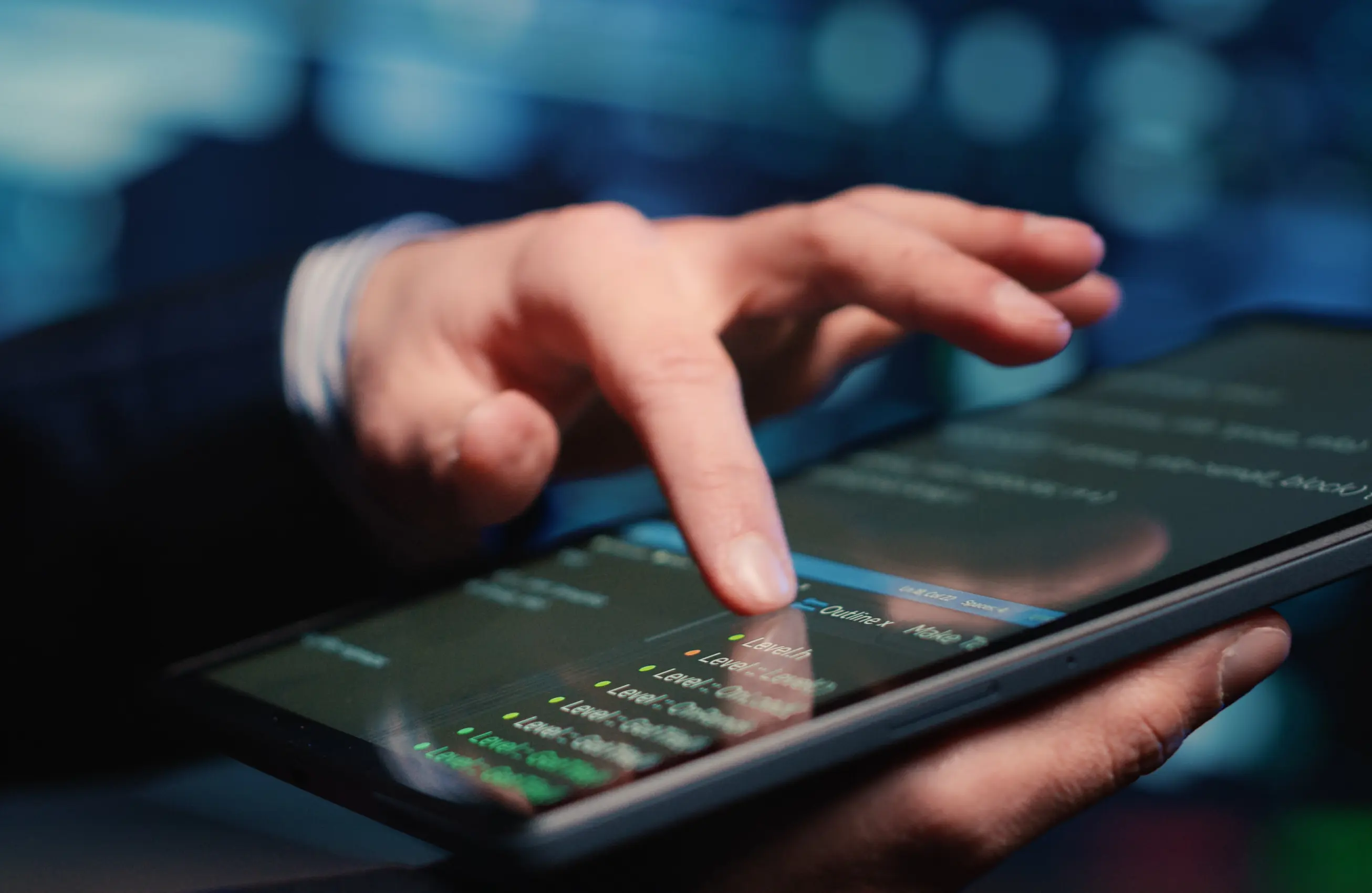 Mão apontando ou tocando a tela de um tablet em um ambiente com iluminação azul, com textos/linhas visíveis na tela.