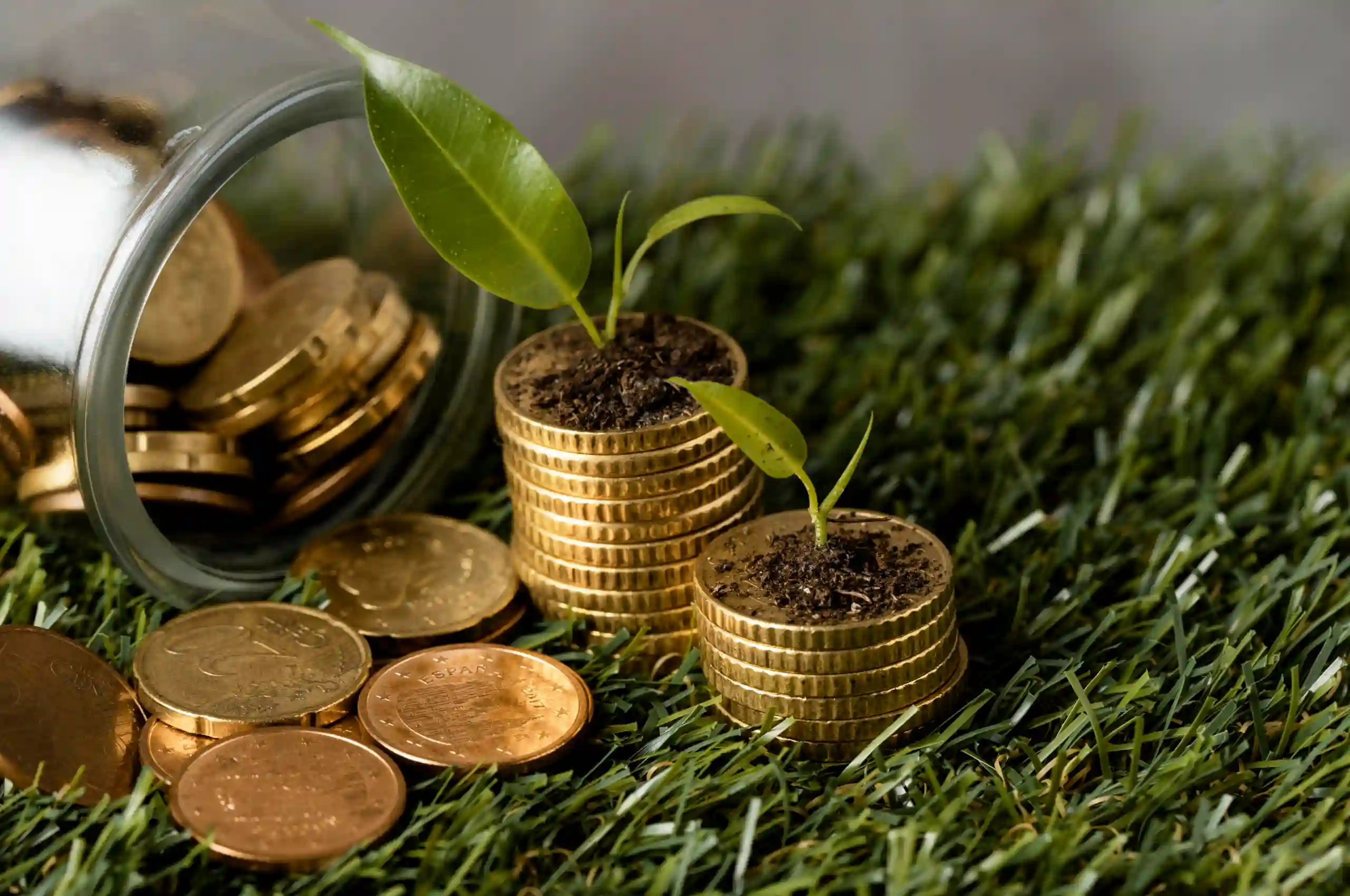 Pilha de moedas douradas sobre grama artificial, com duas mudas verdes plantadas em cima das moedas. Ao lado, um pote de vidro deitado com moedas espalhadas ao redor, representando o conceito de investimento sustentável e crescimento financeiro.