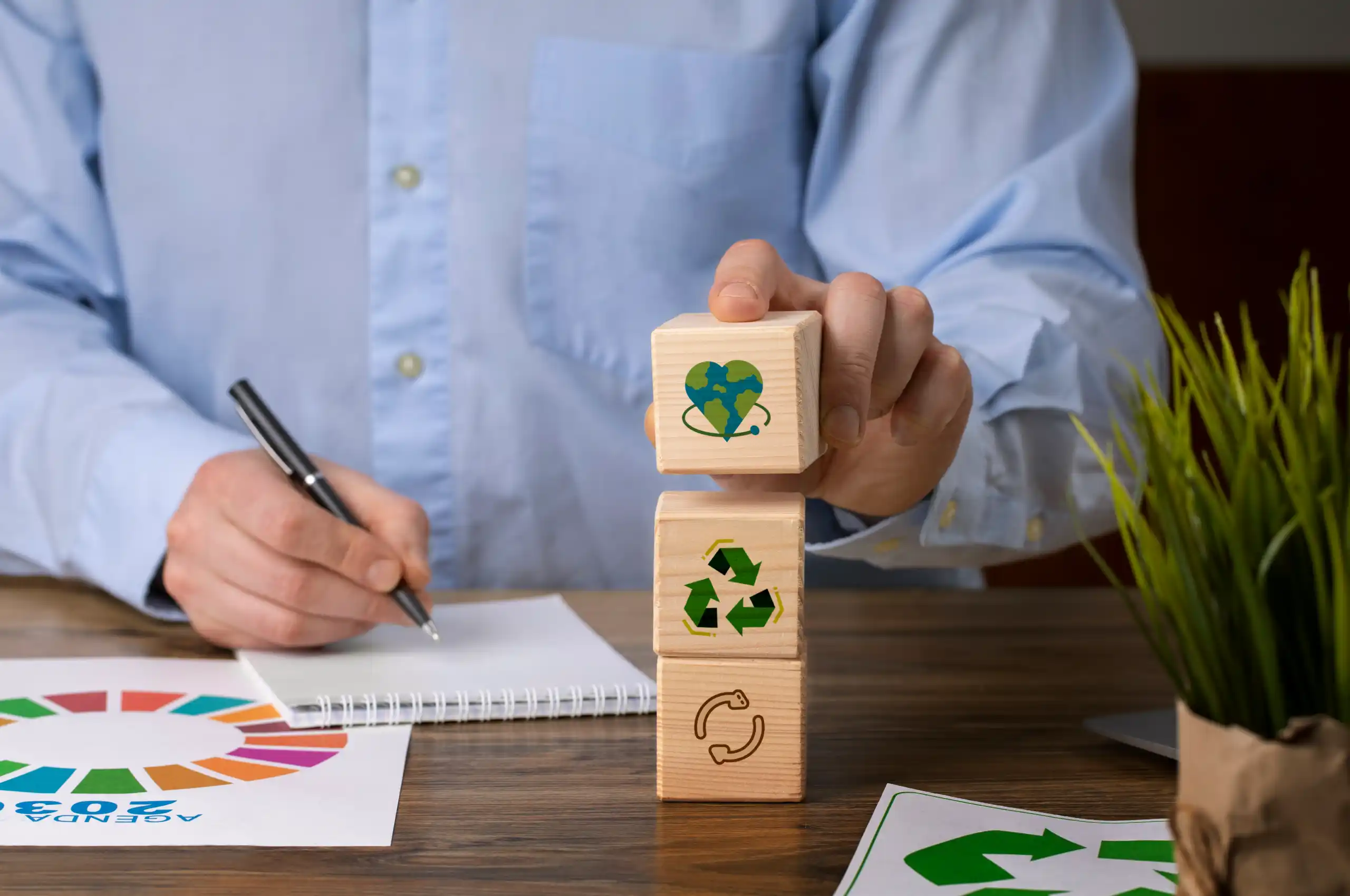 Pessoa com camisa azul clara empilhando blocos de madeira com símbolos de sustentabilidade, como reciclagem e coração com o planeta Terra. Sobre a mesa há um caderno, uma caneta, papéis com ícones de reciclagem e uma planta, representando conceitos de responsabilidade ambiental e desenvolvimento sustentável. 