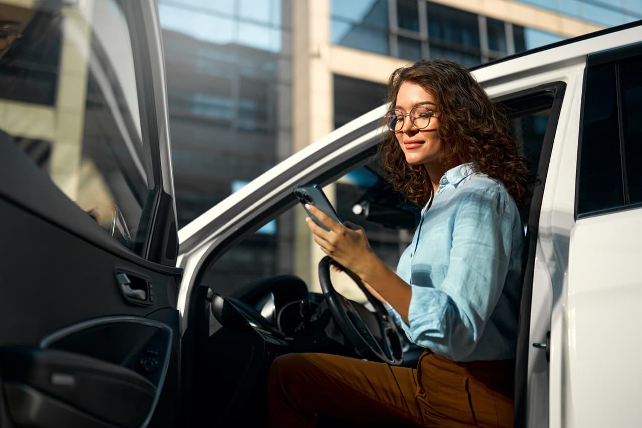 Mulher sentada no carro, com porta aberta mexendo no celular