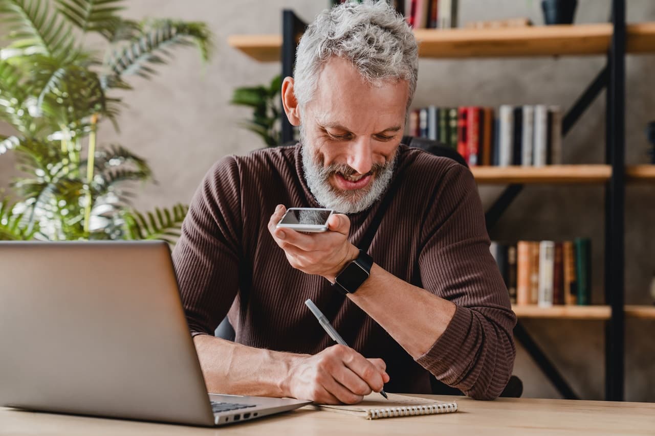 homem sentado em frente a mesa, mexendo no celular, com notebook a frente.