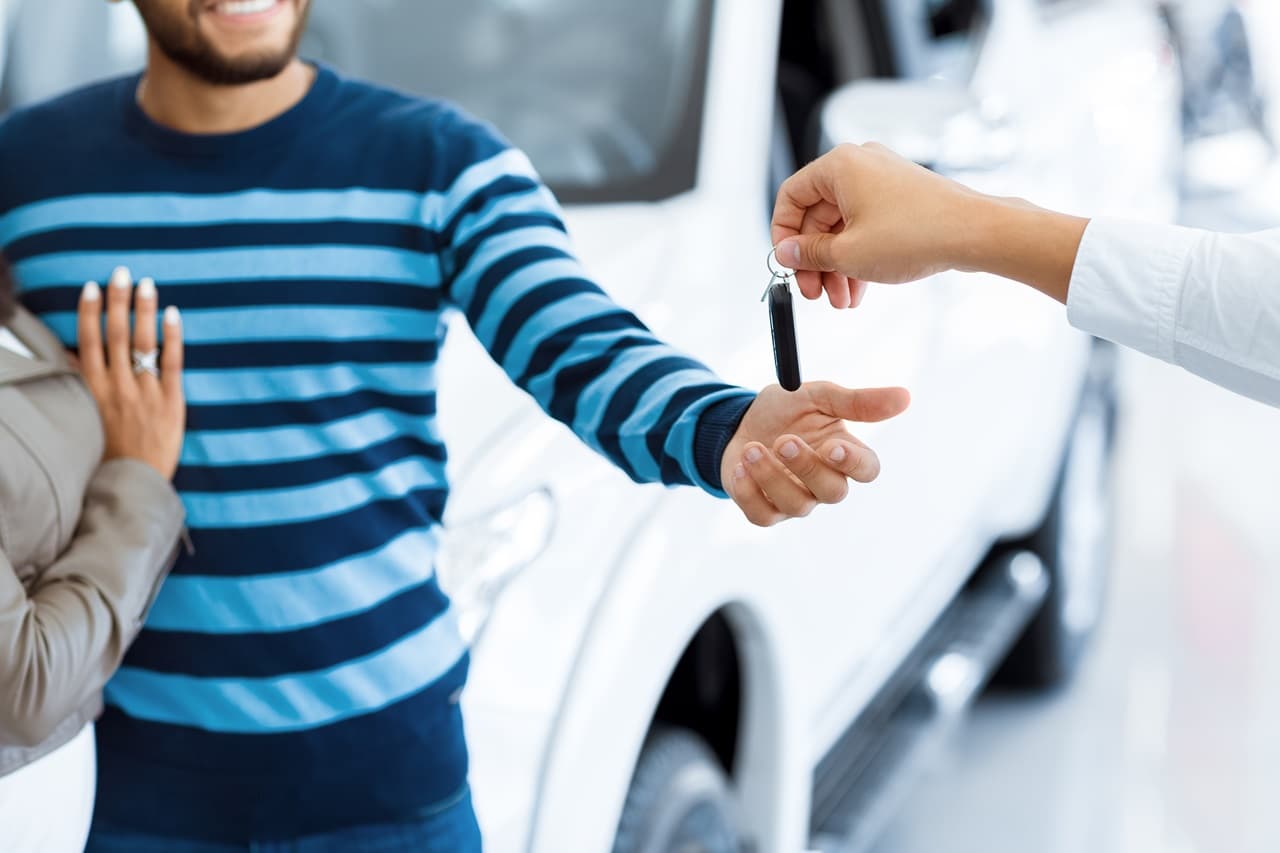 Casal em frente a um carro com as mãos estendidas, pegando chave do carro que está sendo entregue pelo vendedor