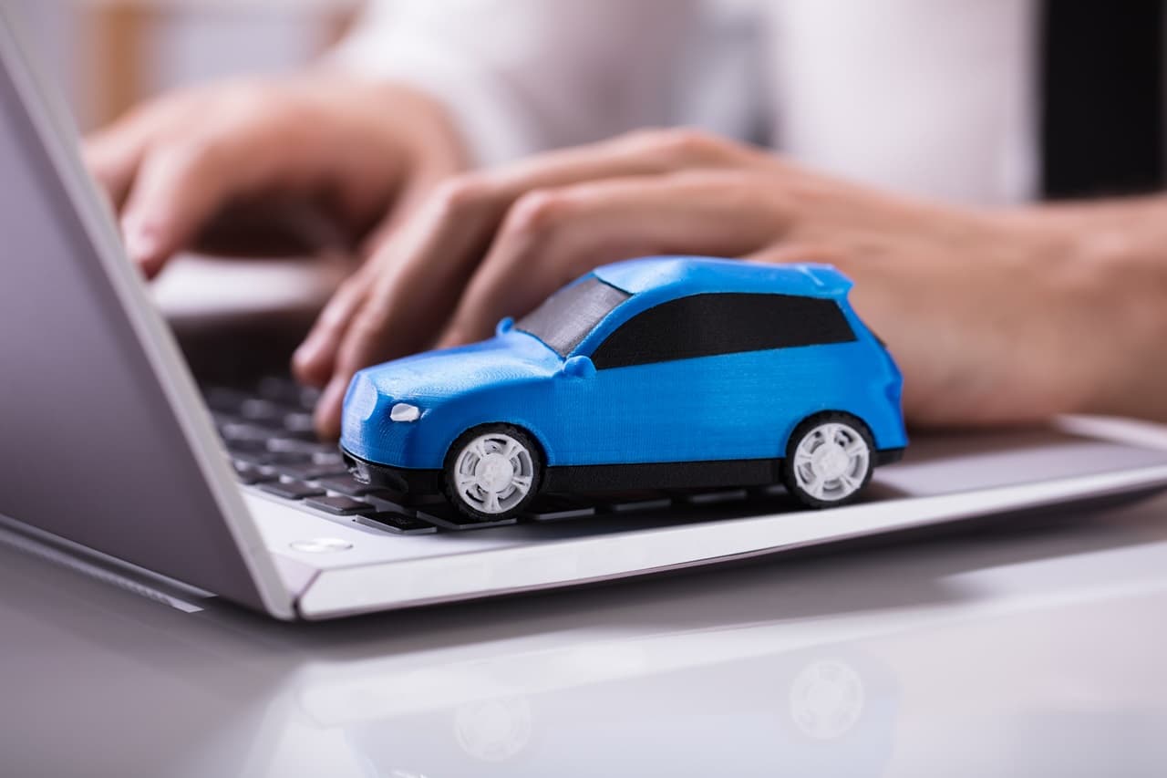 Miniatura de carro azul sobre o teclado de um notebook enquanto uma pessoa digita, representando pesquisa ou planejamento para compra de veículo.