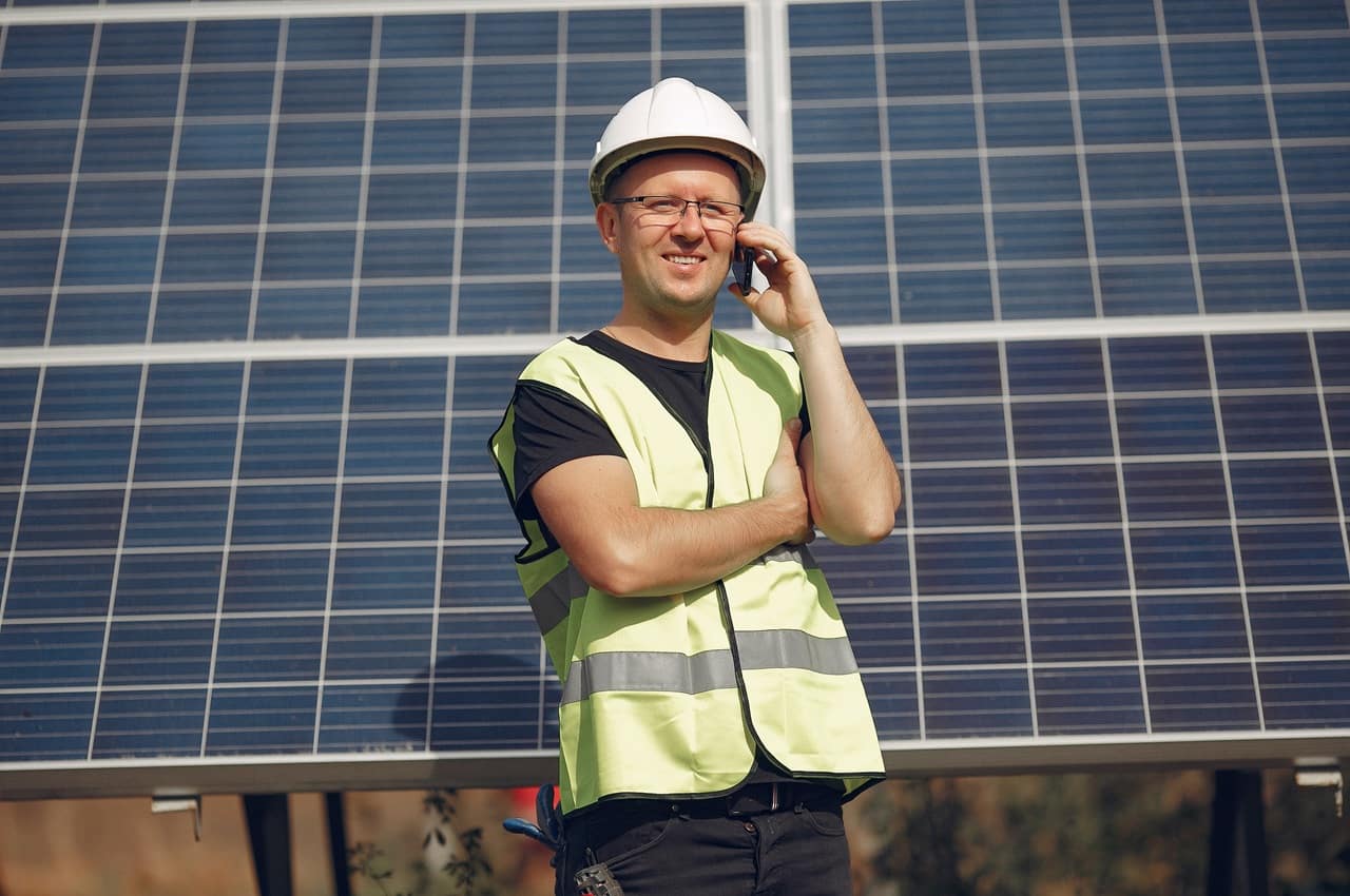 Homem usando capacete branco e colete de segurança amarelo. Ele está em frente a painéis solares instalados e falando ao telefone, com um semblante feliz.