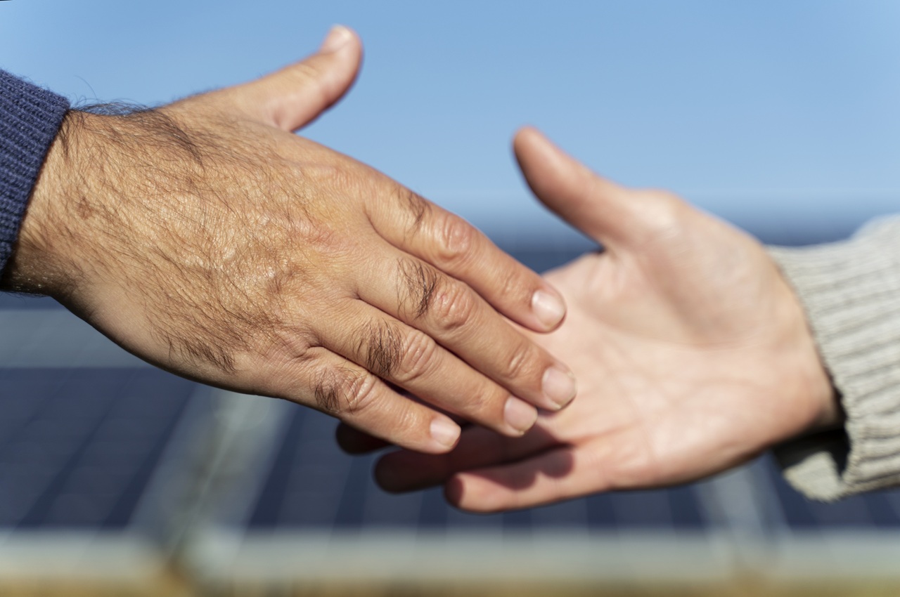 Close-up em um aperto de mãos de duas pessoas, com um painel solar ao fundo, indicando fechamento de parceria com um fornecedor de energia solar.