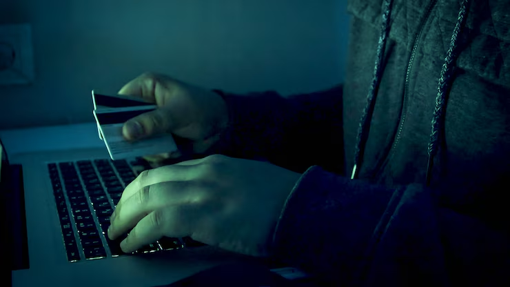 Uma imagem close-up de uma pessoa utilizando luvas pretas enquanto digita códigos em um notebook e segura um cartão de crédito. 