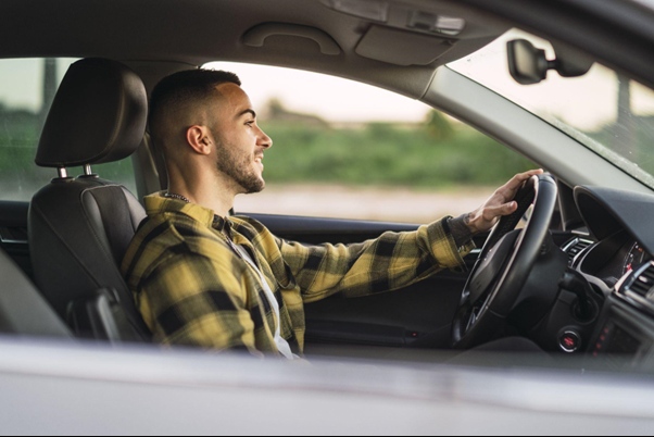 Vista interna de um jovem motorista sorridente ao volante de um carro em movimento. Ele usa uma camisa de flanela xadrez amarela e tem tatuagens no braço, aparentando estar aproveitando o trajeto.