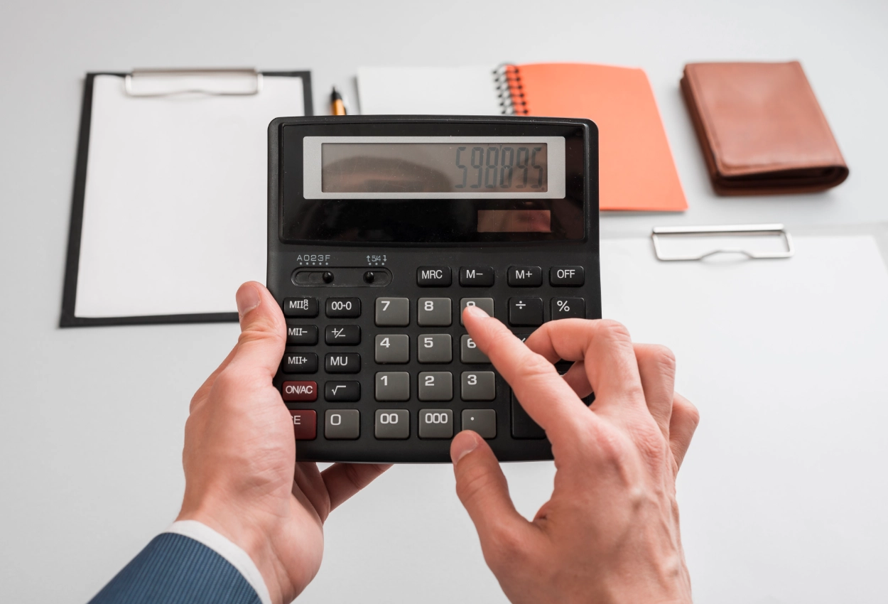 Close-up de mãos masculinas segurando calculadora financeira. Ao fundo, há uma mesa branca com pranchetas e cadernetas.