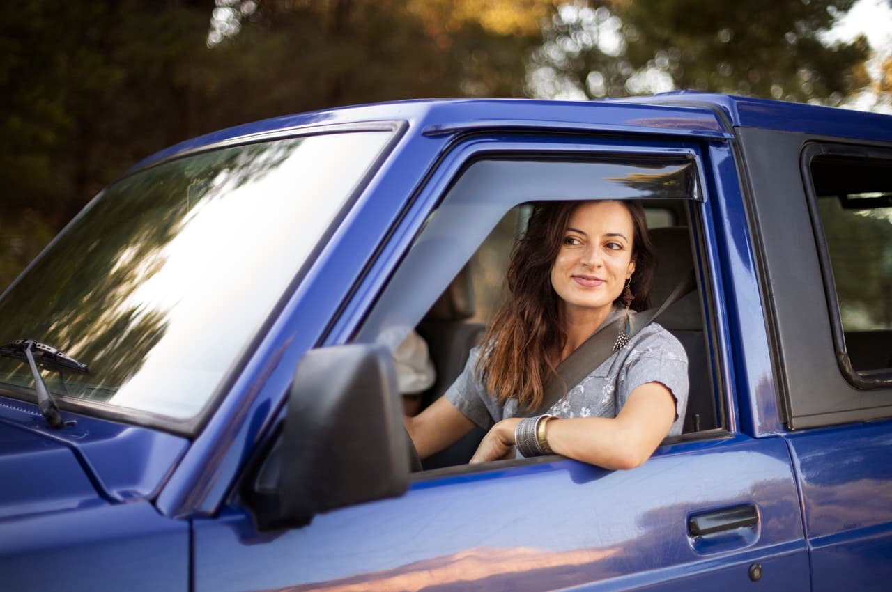 Mulher sorridente dirigindo carro usado azul e olhando pela janela. A mulher tem cabelos castanhos longos, usa cinto de segurança e veste camisa de magas curtas cinzas