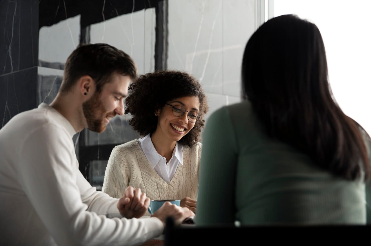 Três pessoas conversando enquanto estão sentadas à mesa, aparentemente em uma reunião de trabalho. O foco está em uma mulher de óculos e cabelos crespos sorrindo para os seus colegas.