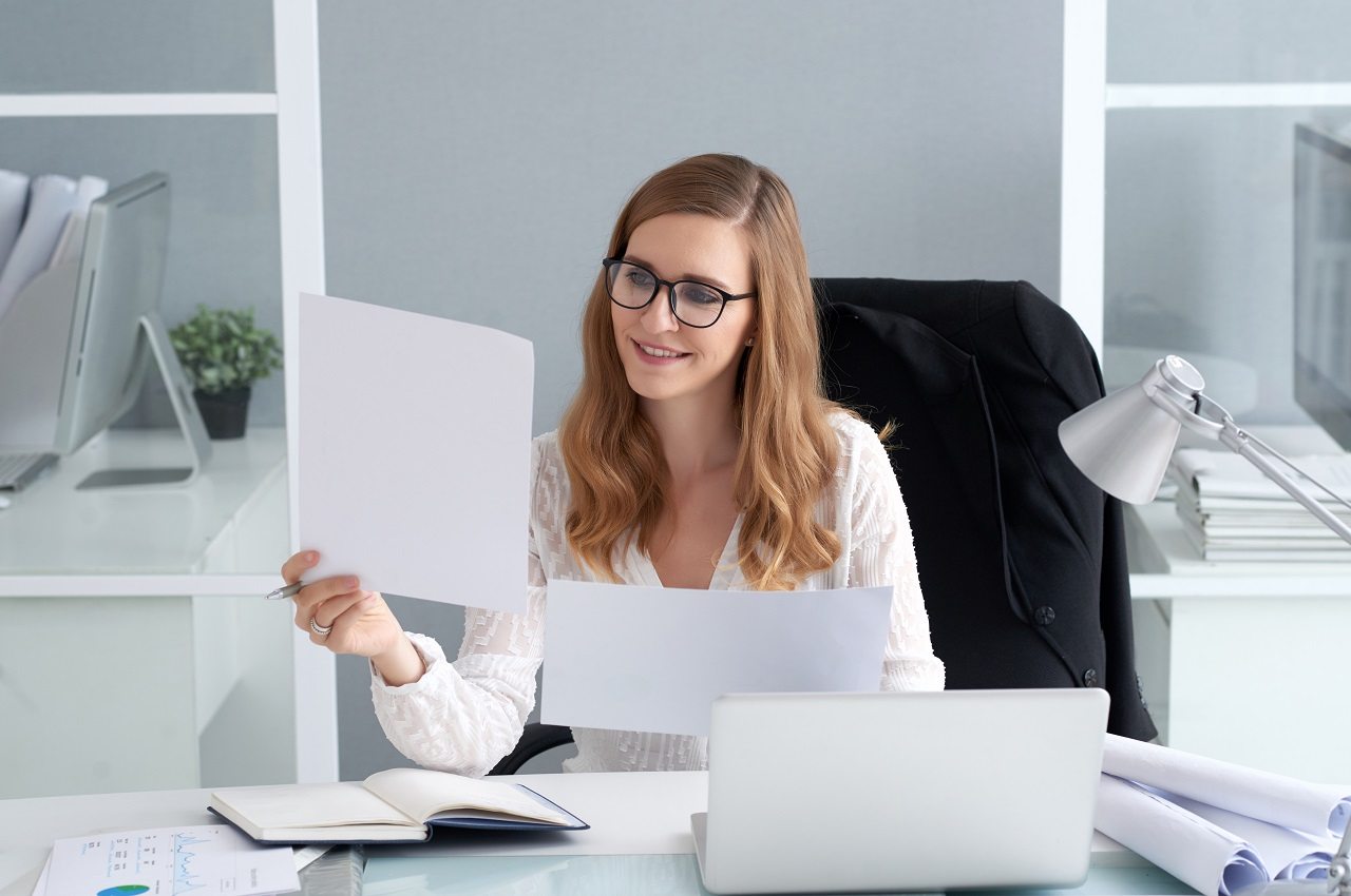 Mulher sorridente de cabelos longos loiros sentada em uma mesa de escritório e segurando folhas e caderno aberto ao lado de um laptop.