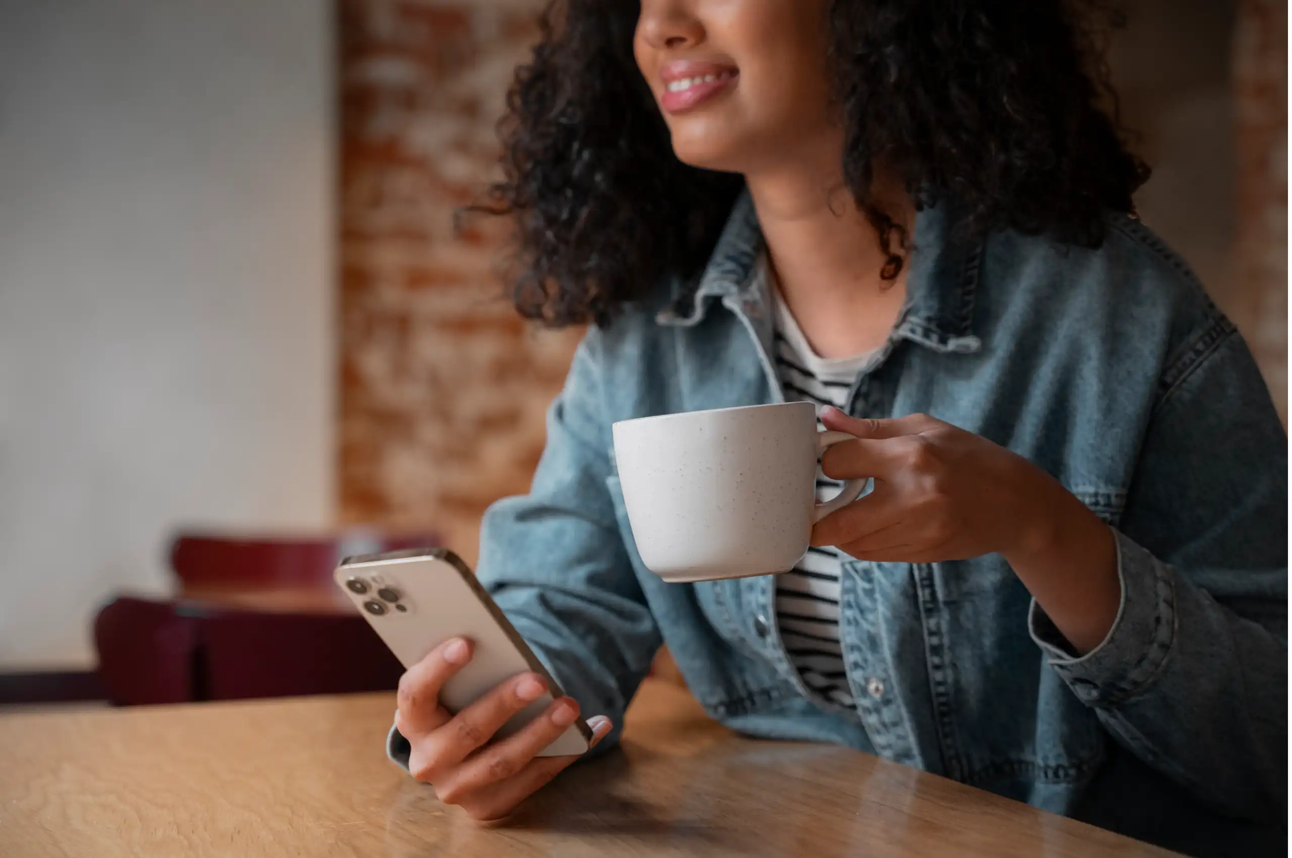 Uma mulher sentada em uma mesa de madeira. Ela está segurando uma caneca com a mão esquerda e um smartphone na mão direita.