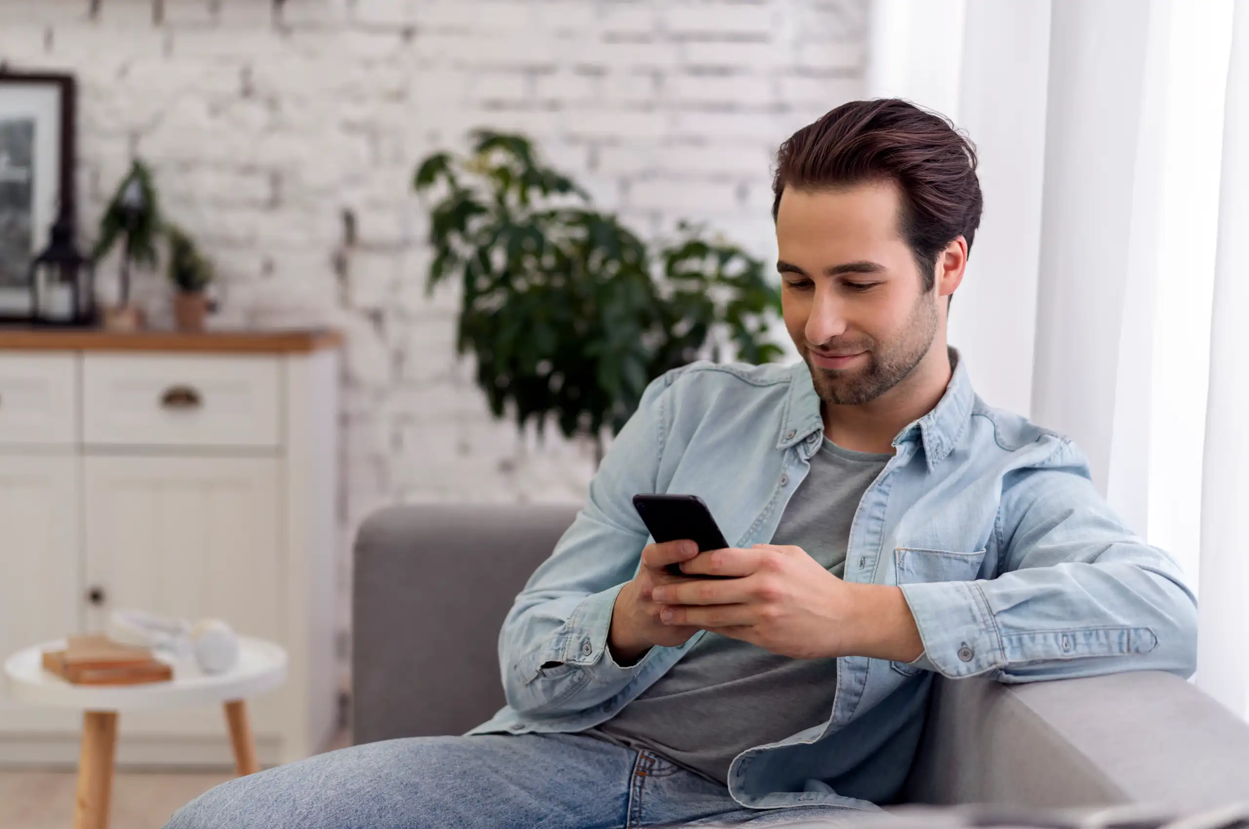 Um homem sentado em um sofá de uma sala de estar enquanto utiliza um smartphone sorrindo.