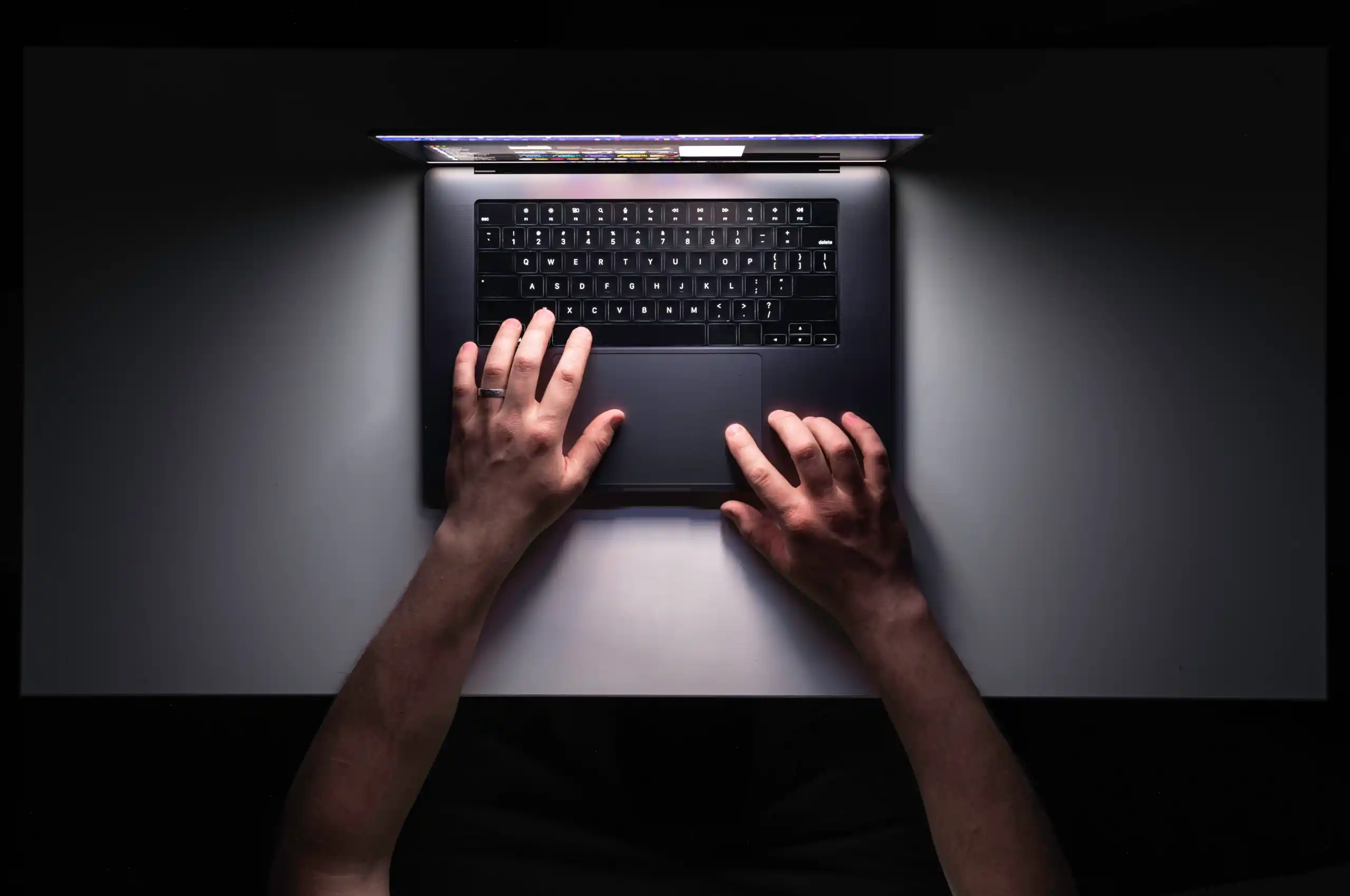Imagem mostrando a visão de cima de uma mesa de trabalho, o ambiente está escuro e a luz da tala do computador ilumina o local enquanto a mão de um homem trabalha no aparelho.