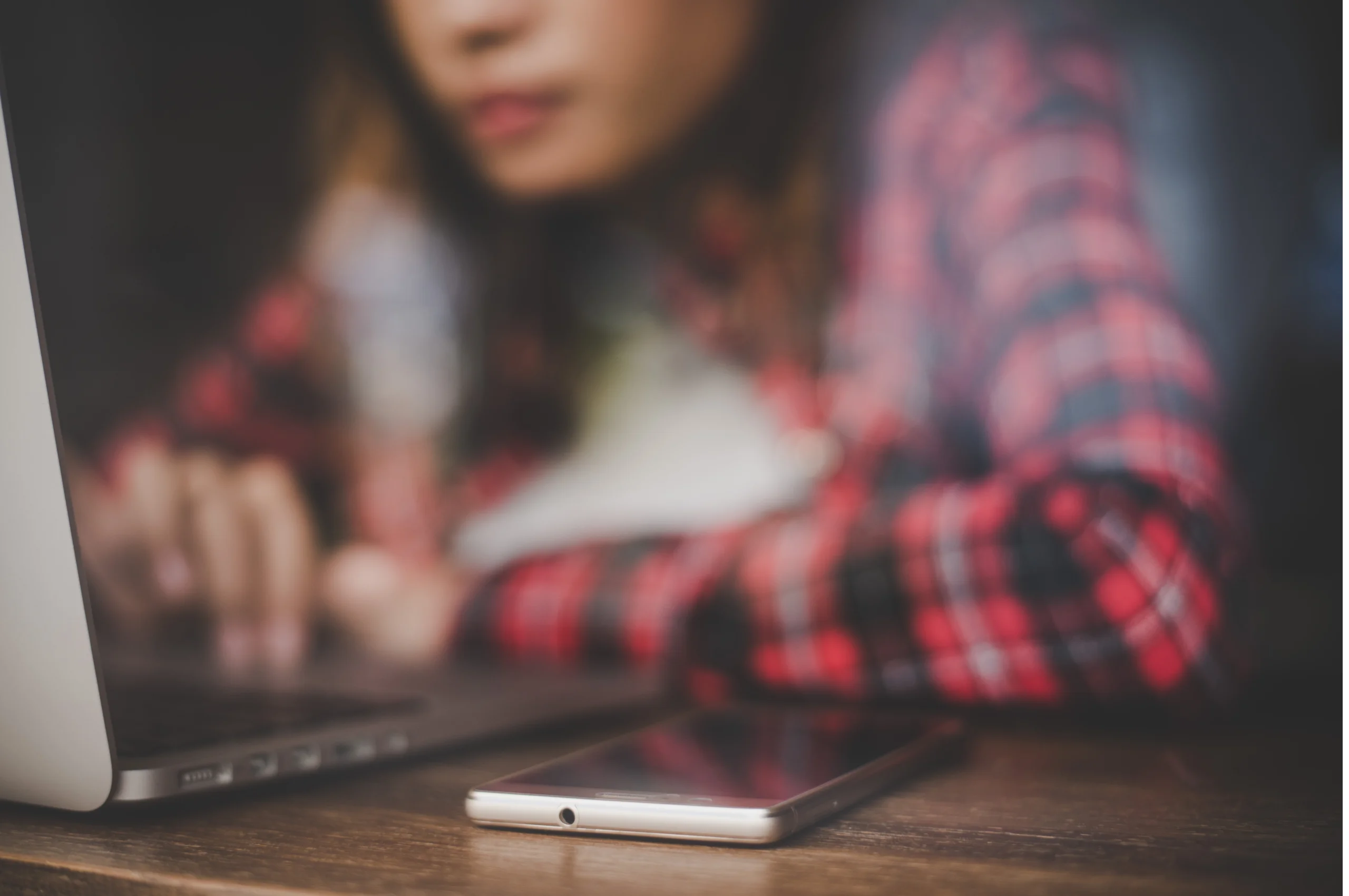Pessoa jovem em camiseta xadrez vermelha usando um laptop, com celular colocado na mesa ao lado; foco suave na tela.