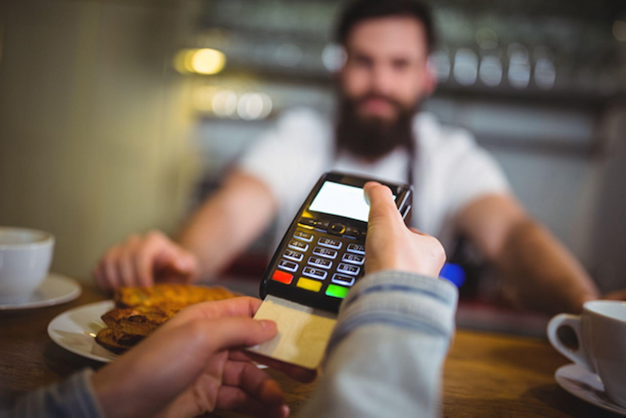 Uma máquina de cartão é segurada por uma mão, enquanto o cartão é inserido para pagamento. Ao fundo, a imagem mostra xícaras de café, um doce e o atendente da cafeteria.