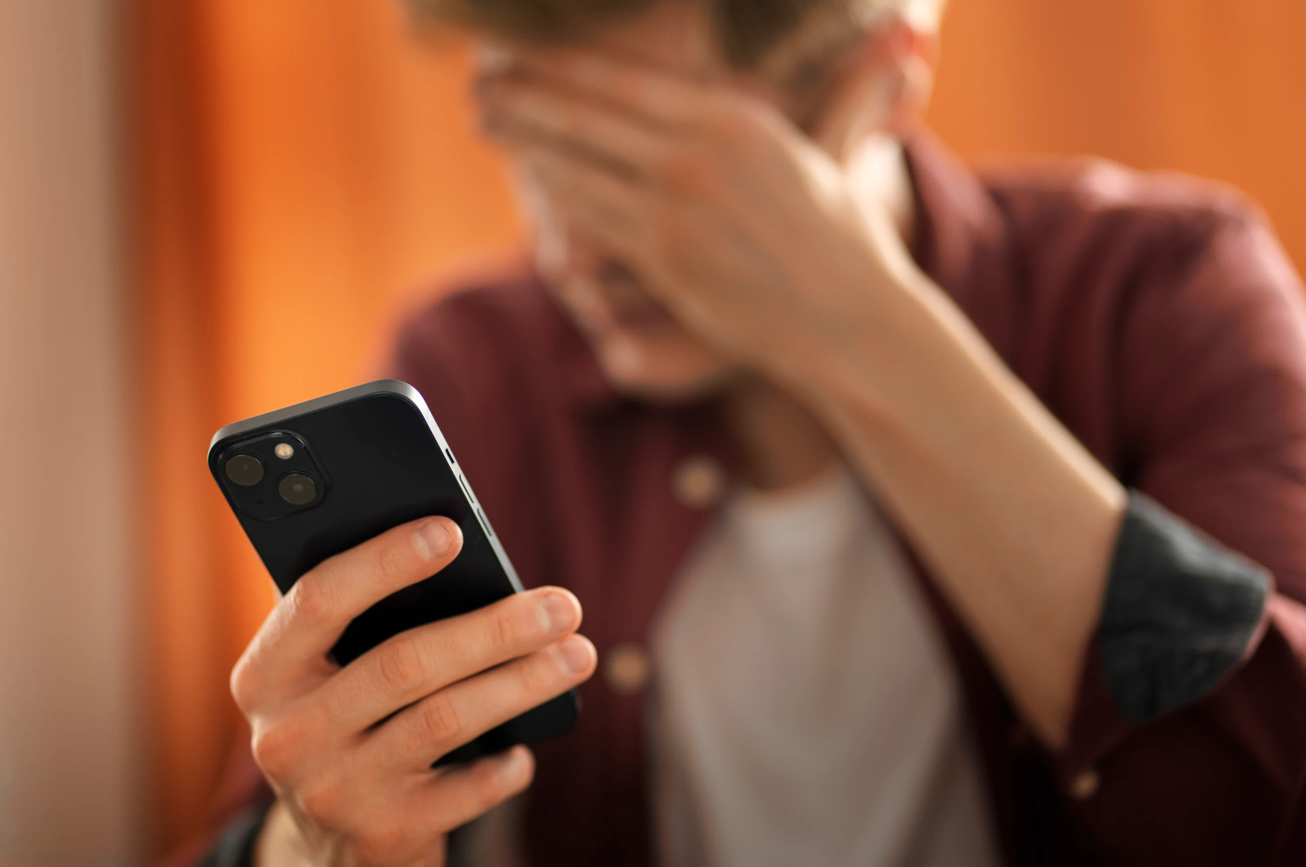 Pessoa segurando um celular preto com uma das mãos, enquanto a outra mão cobre parte do rosto; fundo com cortinas laranjas desfocado.