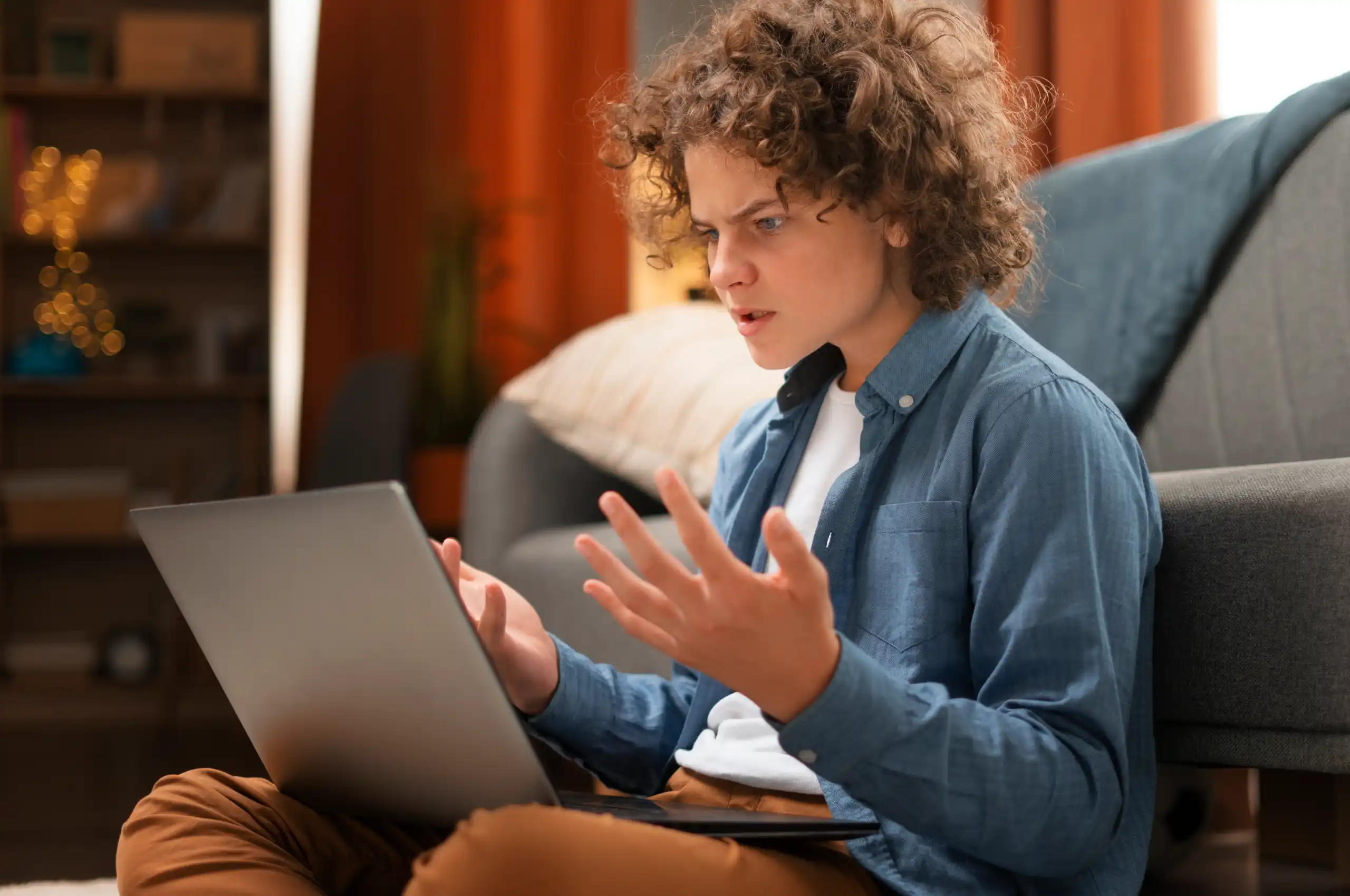 Jovem sentado no chão da sala, usando laptop em geral aberto, expressão de surpresa enquanto gesticula com as mãos; ao fundo há sofá e estante. 