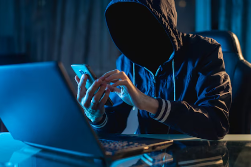Um homem com um capuz escondendo o rosto em uma sala escura. Ele está sentado na frente de um notebook enquanto utiliza um smartphone.