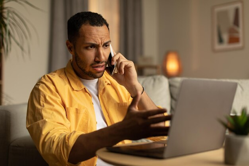 Um homem sentado sobre uma mesa com um notebook, enquanto fala algo em uma ligação no celular e aponta para a tela do aparelho.