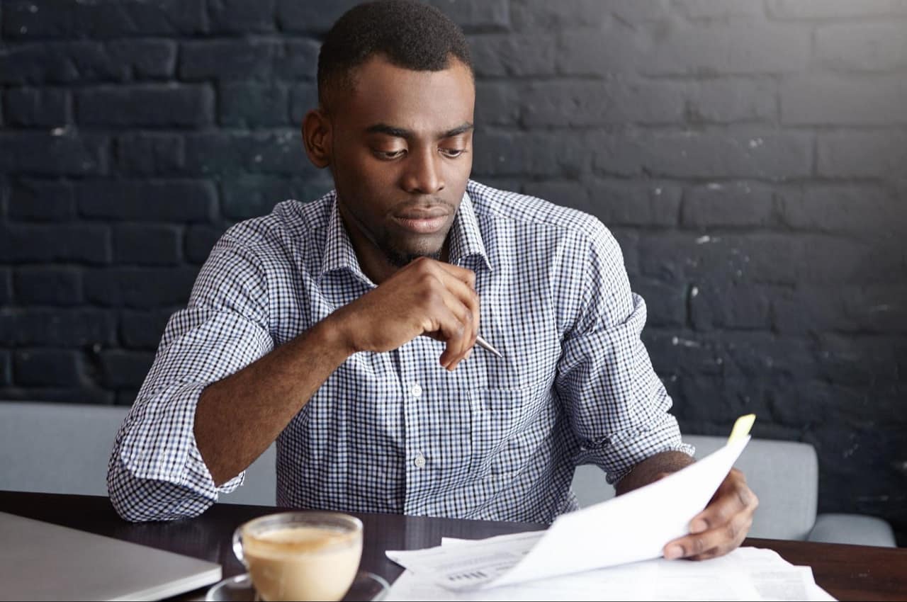 Homem com expressão concentrada analisando contrato de venda de veículos e tomando xícara de café que está em cima da mesa.