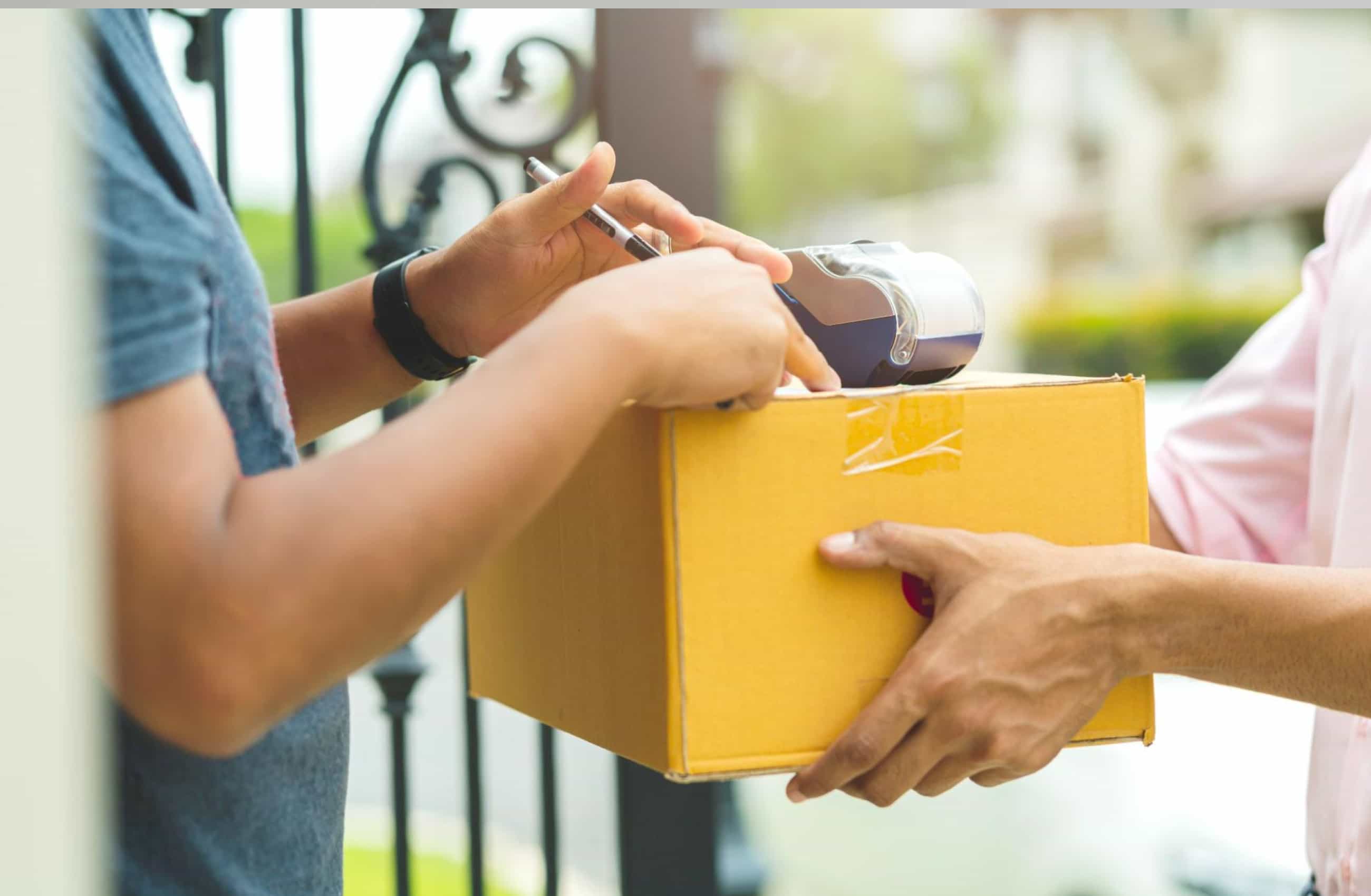 Mão de um entregador segurando uma caixa de papelão amarela enquanto o cliente utiliza uma maquininha de cartão para realizar o pagamento, momento em que pode ocorrer o golpe da entrega falsa.