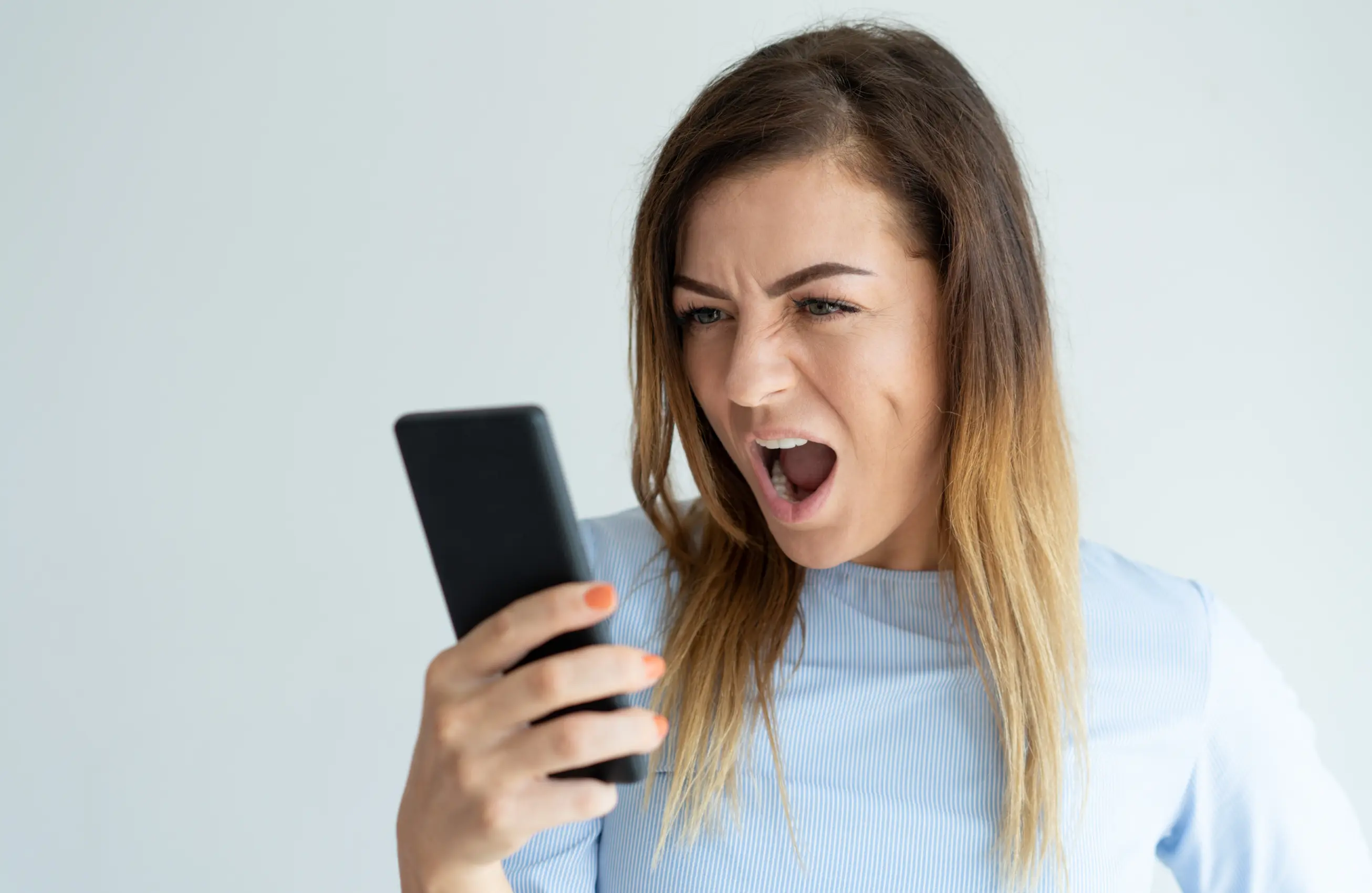 Mulher olhando para o celular com expressão de raiva ou surpresa, boca aberta e sobrancelhas franzidas, segurando o aparelho com a mão direita.&nbsp;