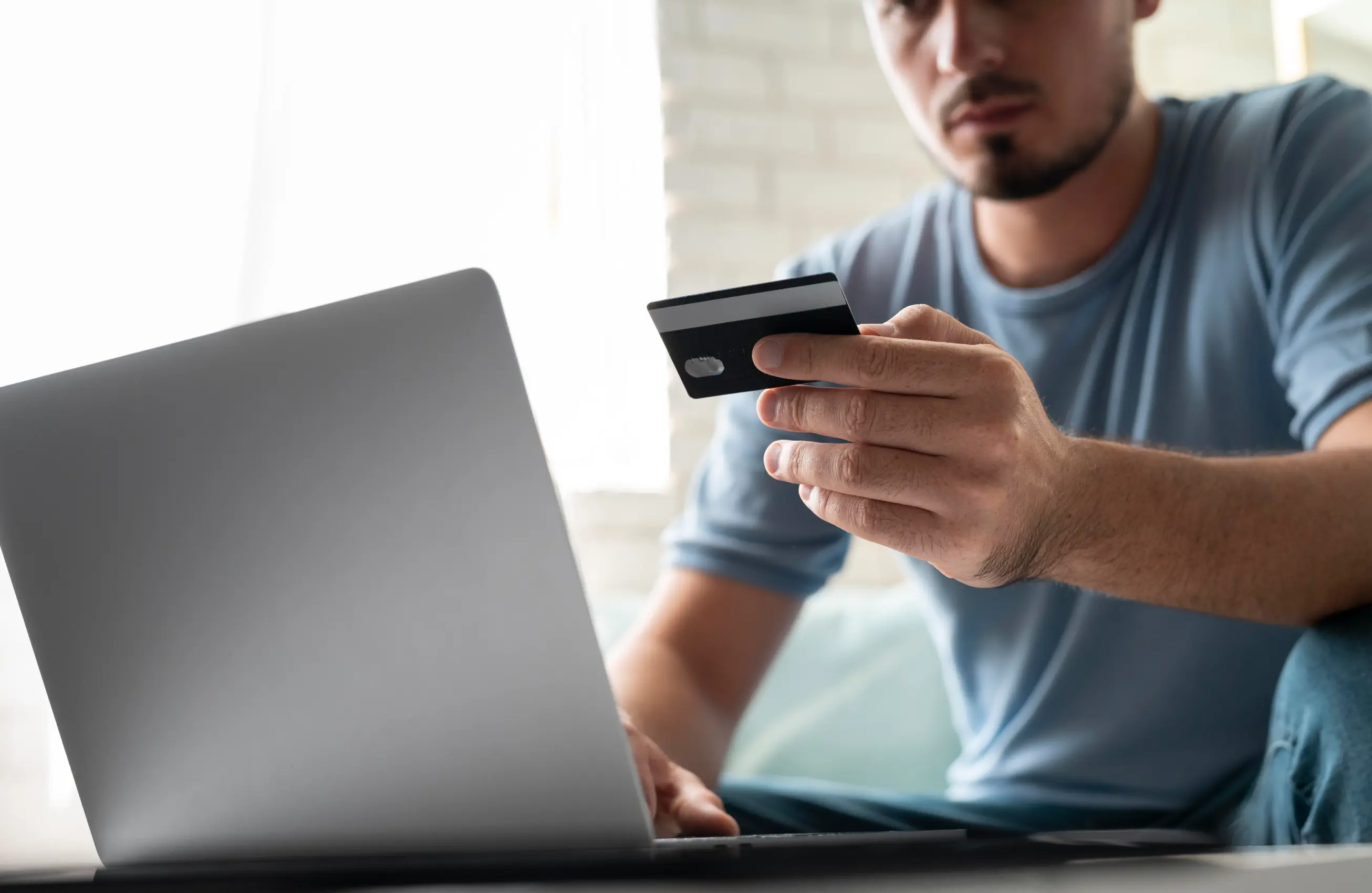 Homem segurando um cartão de crédito na mão direita e usando um laptop, em ambiente interno.