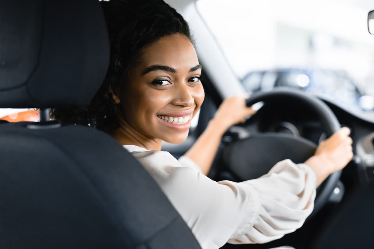 Pessoa sentada no banco do motorista de um carro, com as mãos no volante, olhando em direção à câmera.