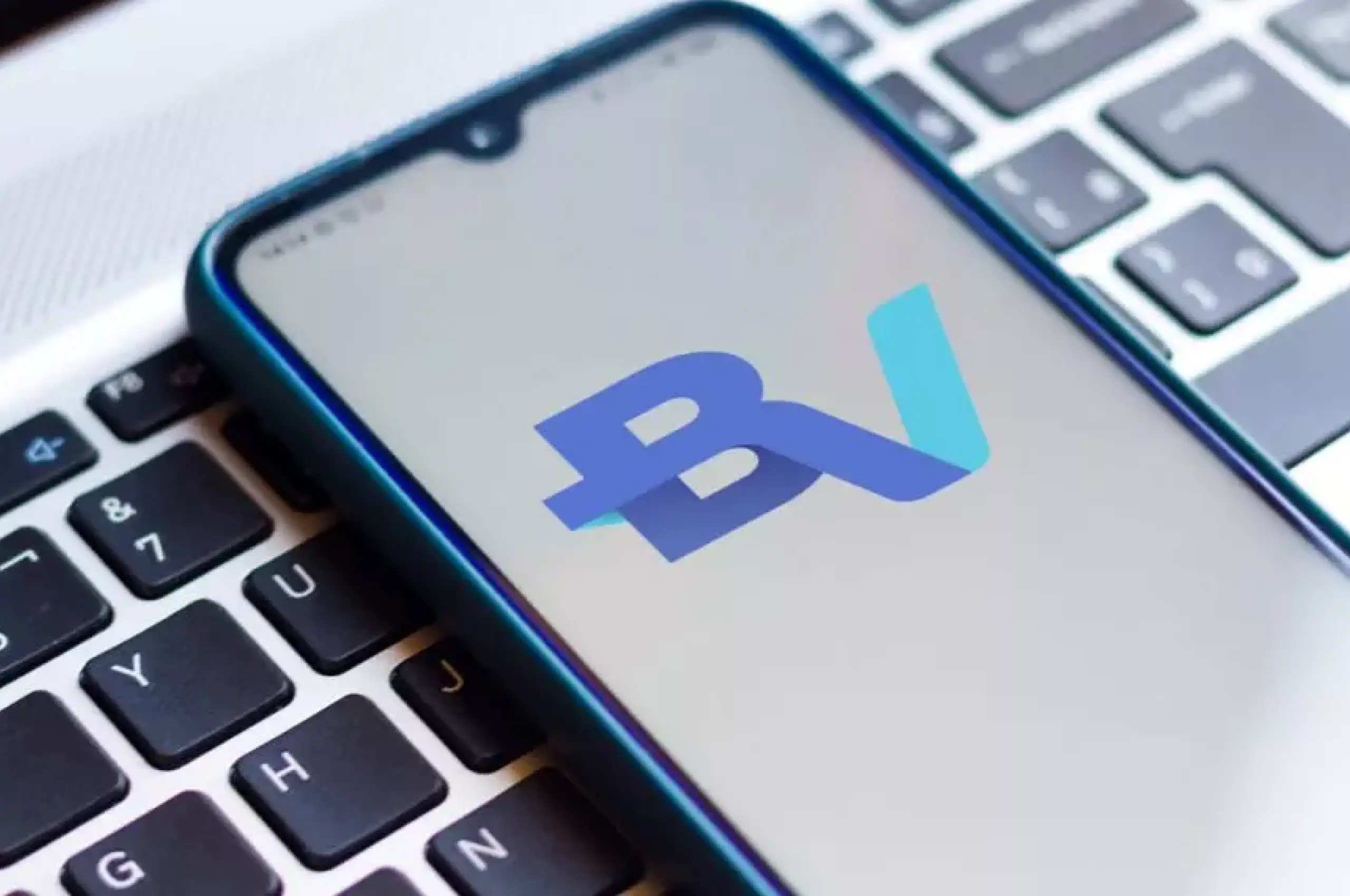 Um smartphone apoiado em um teclado do notebook. A tela do aparelho exibe a logo do BV sobre um fundo branco. 