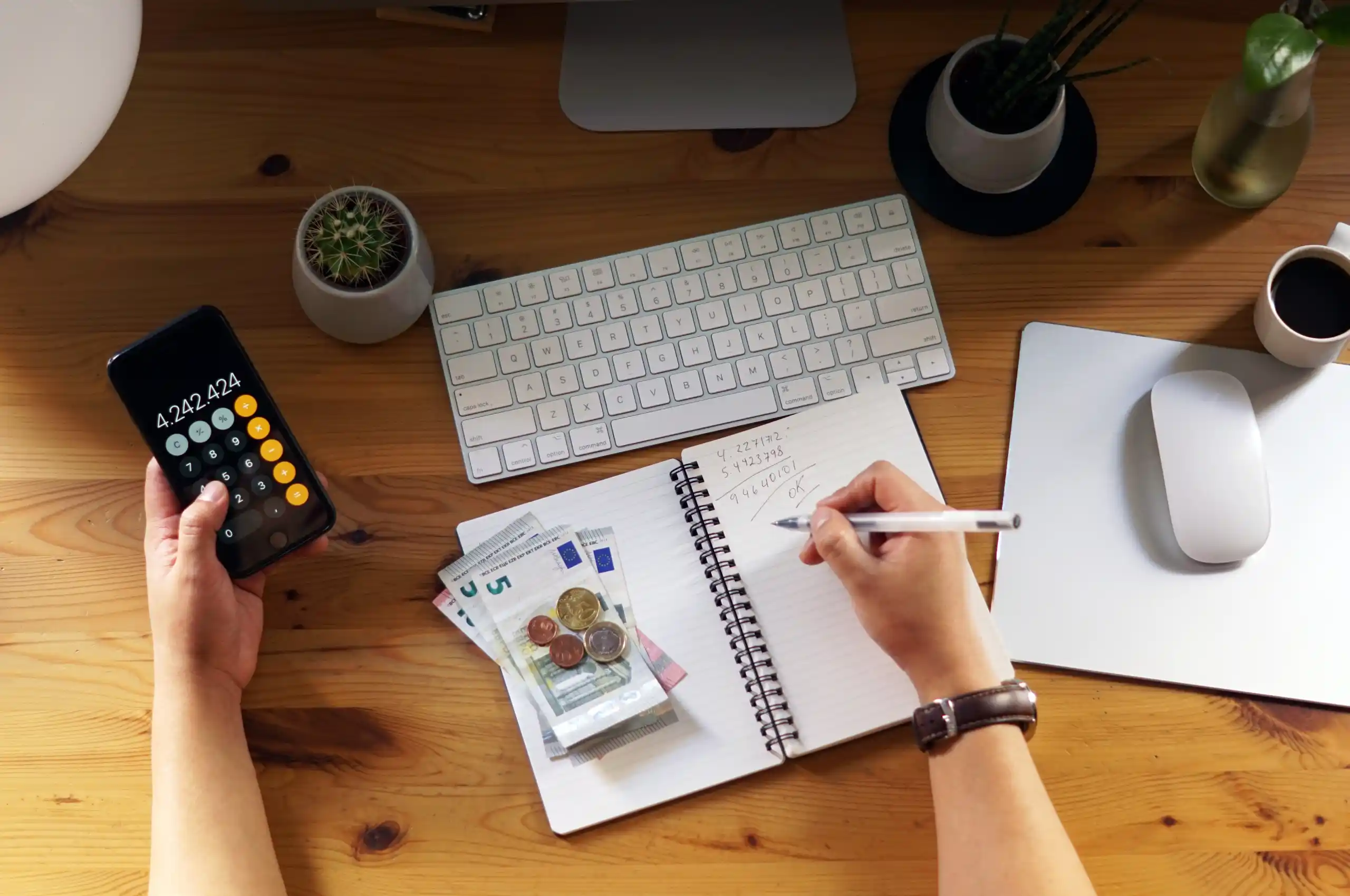 Pessoa planejando compartilhar o histórico financeiro em mesa de trabalho: uma mão segura celular com calculadora aberta e a outra anota valores em caderno ao lado de notas, moedas, teclado, mouse, plantas e uma xícara.