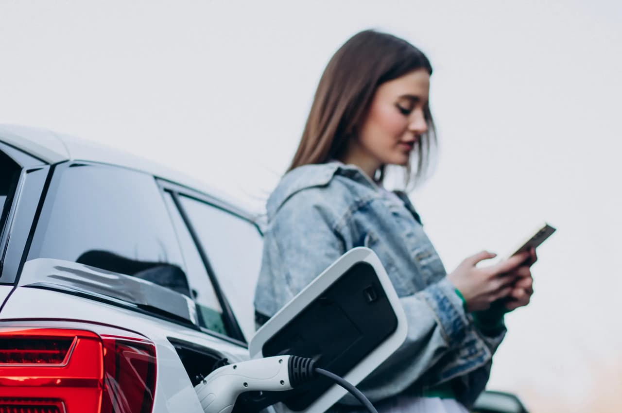 Mulher ao lado de um carro elétrico branco conectado ao carregador, enquanto utiliza o celular. Ela veste uma jaqueta jeans e está em um ambiente externo.