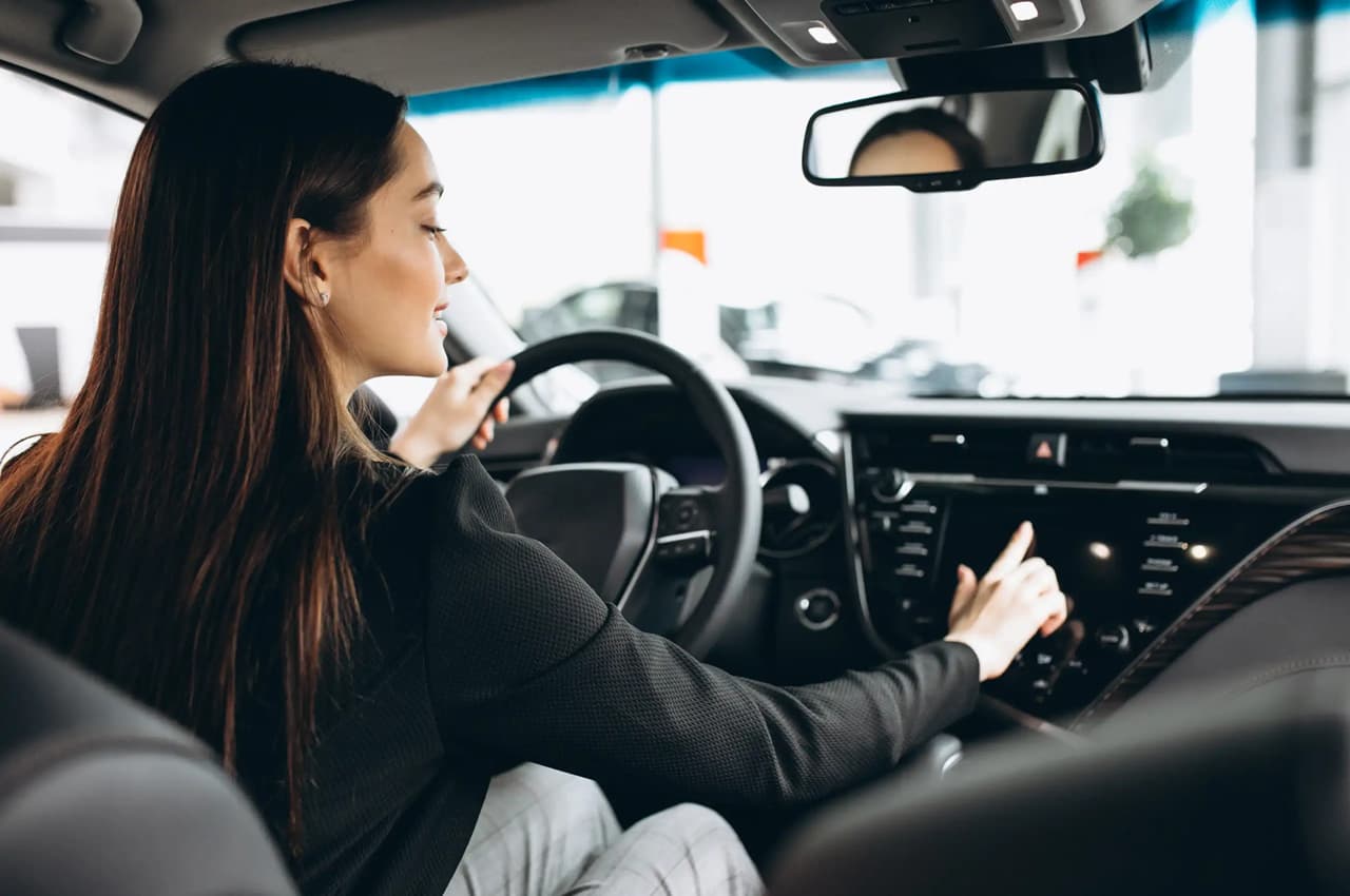 Mulher jovem no banco do motorista de um carro moderno, testando o painel multimídia em uma concessionária. Ela veste roupas sociais e demonstra interesse na tecnologia do veículo.