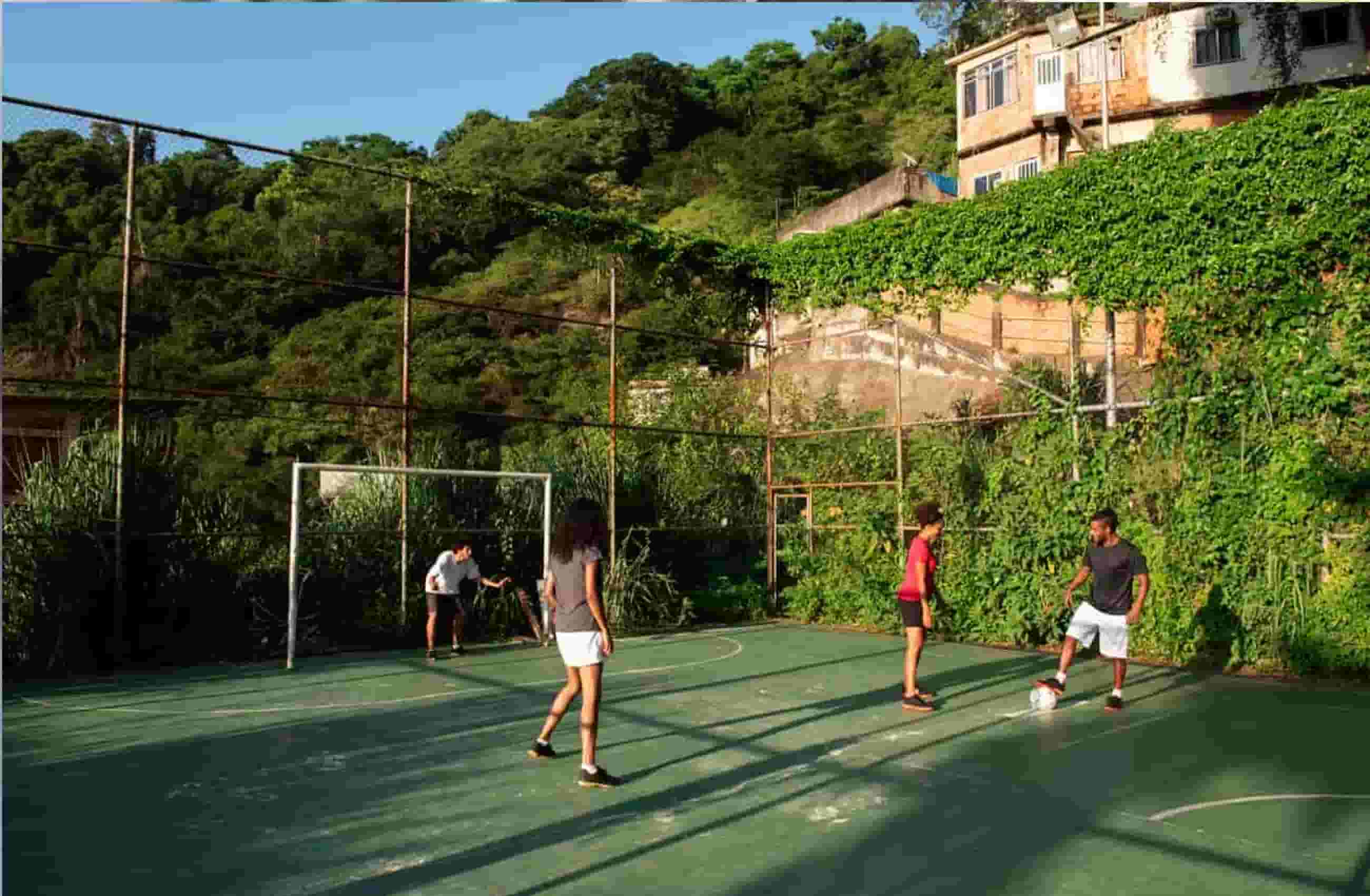 Quatro pessoas jogando futebol em um campo. Uma está com a bola nos pés, outro está no gol e duas pessoas estão esperando para jogar.