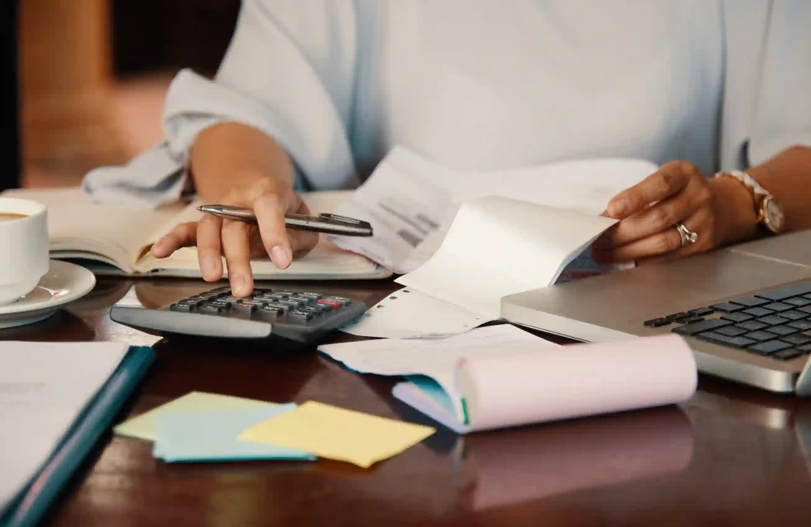 Visão de uma mesa de escritório com papéis, calculadora e um notebook. Somente as mãos de uma mulher estão visíveis enquanto ela realiza contas.