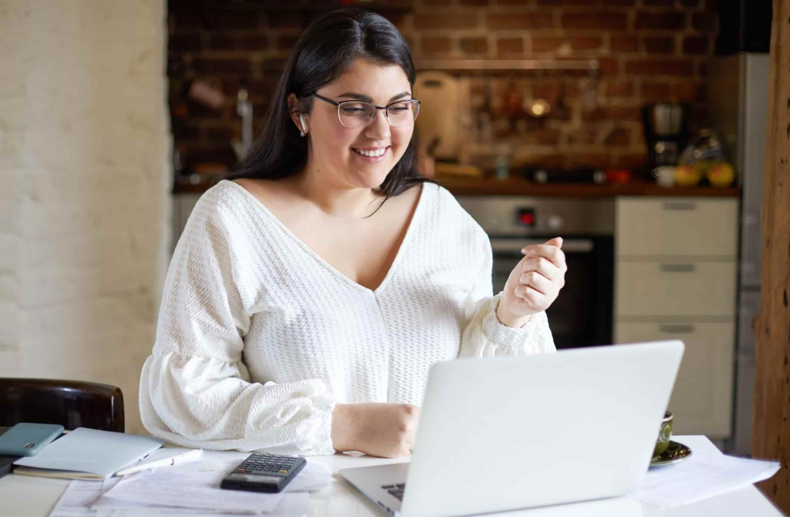 Uma mulher jovem, de óculos e blusa branca, sorri enquanto utiliza um notebook em uma mesa organizada com calculadora e documentos.