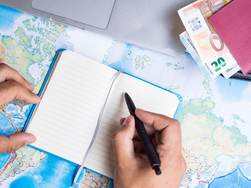 Uma pessoa escrevendo em uma caderneta aberta sobre um mapa mundial. Ao canto é possível observar um notebook e uma carteira com dinheiro físico. 