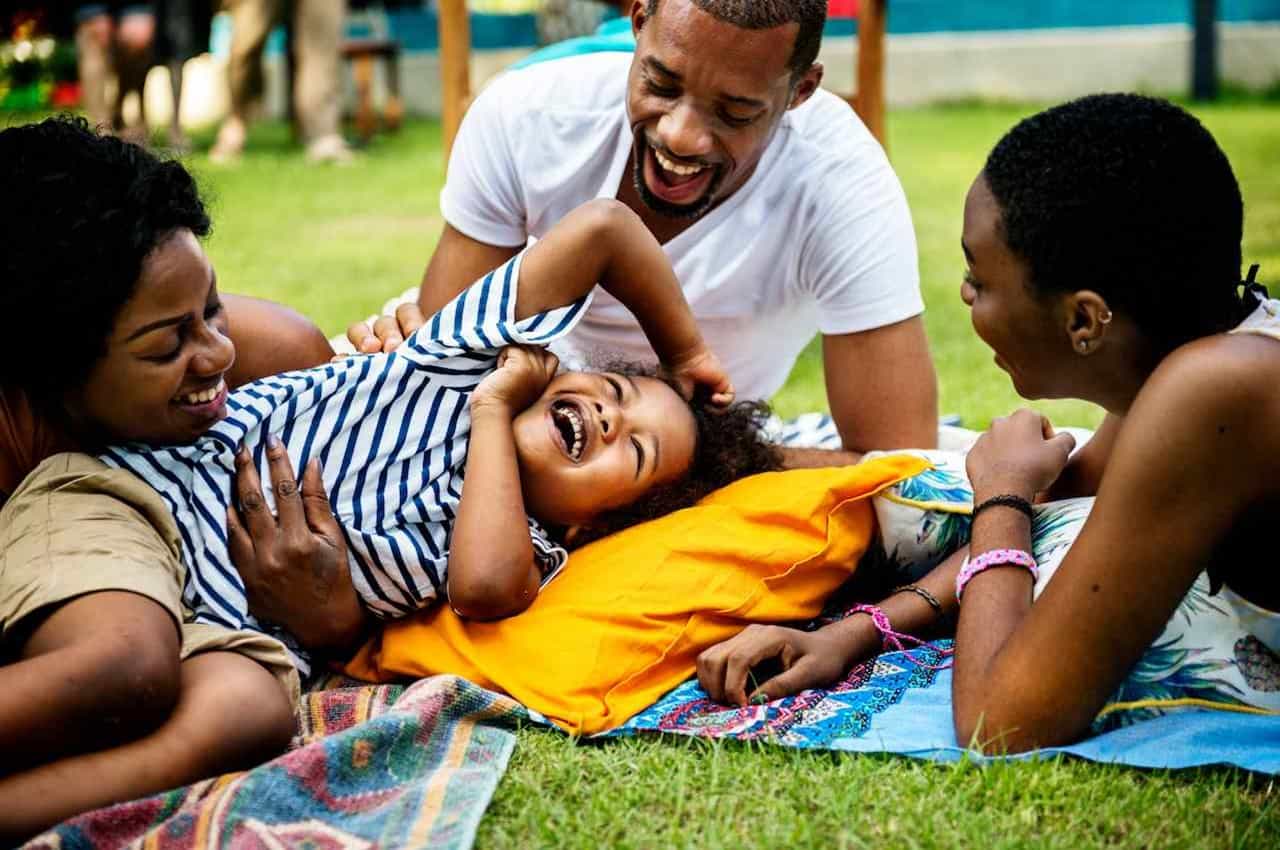 Criança sorridente brincando com seus familiares na grama de uma praça arborizada. Na imagem, tem duas mulheres e um homem que fazem cosquinhas na criança.