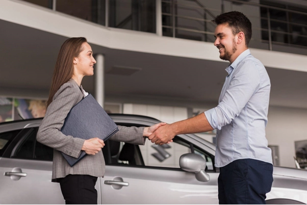 Duas pessoas em frente a um carro branco, apertando as mãos. A mulher segura uma pasta ou caderno, e ambos sorriem, sugerindo acordo concluído.