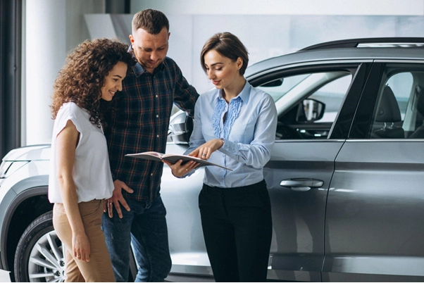 Três pessoas em concessionária automotiva observando um catálogo ou documento aberto, com um carro ao fundo. Uma mulher aponta para o material enquanto os dois colegas ou clientes olham atentamente.