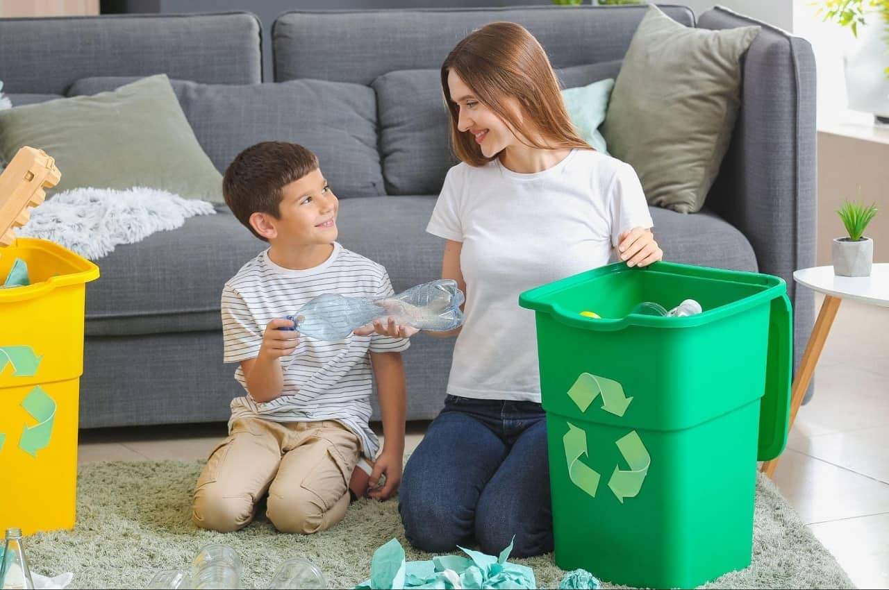Mulher e criança sorridentes sentados no tapete da salar de estar e reciclando garrafas PET e papéis nos lixos adequados.