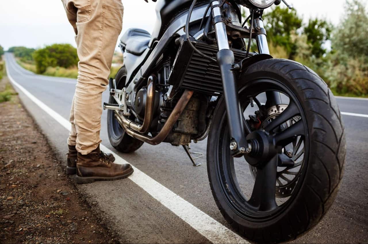 Pernas de uma pessoa em pé, ao lado de uma moto preta estacionada à beira da estrada.