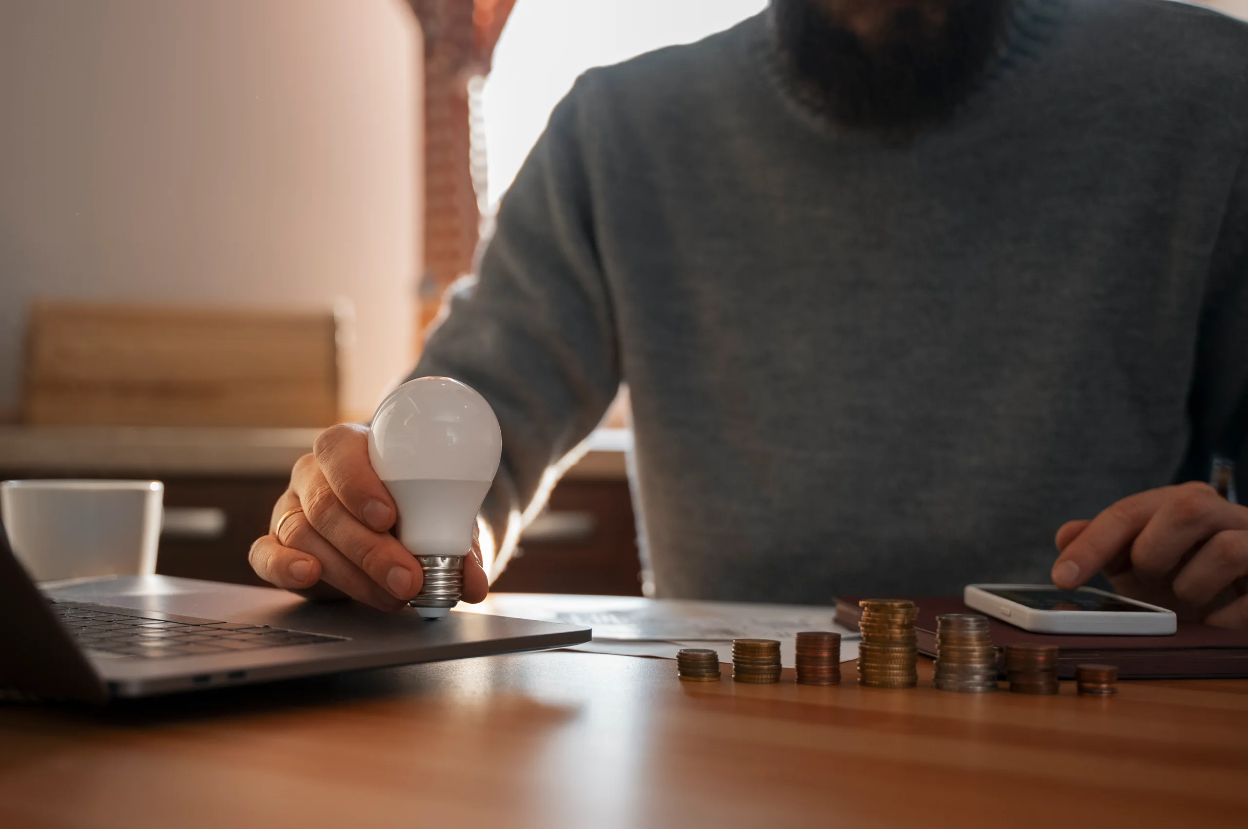 Um homem sentado em uma mesa de madeira enquanto faz contas em um smartphone e segura uma lâmpada, simbolizando uma ideia. Na mesa, é possível observar pilhas de moedas e um notebook. Seu rosto não está visível na foto.