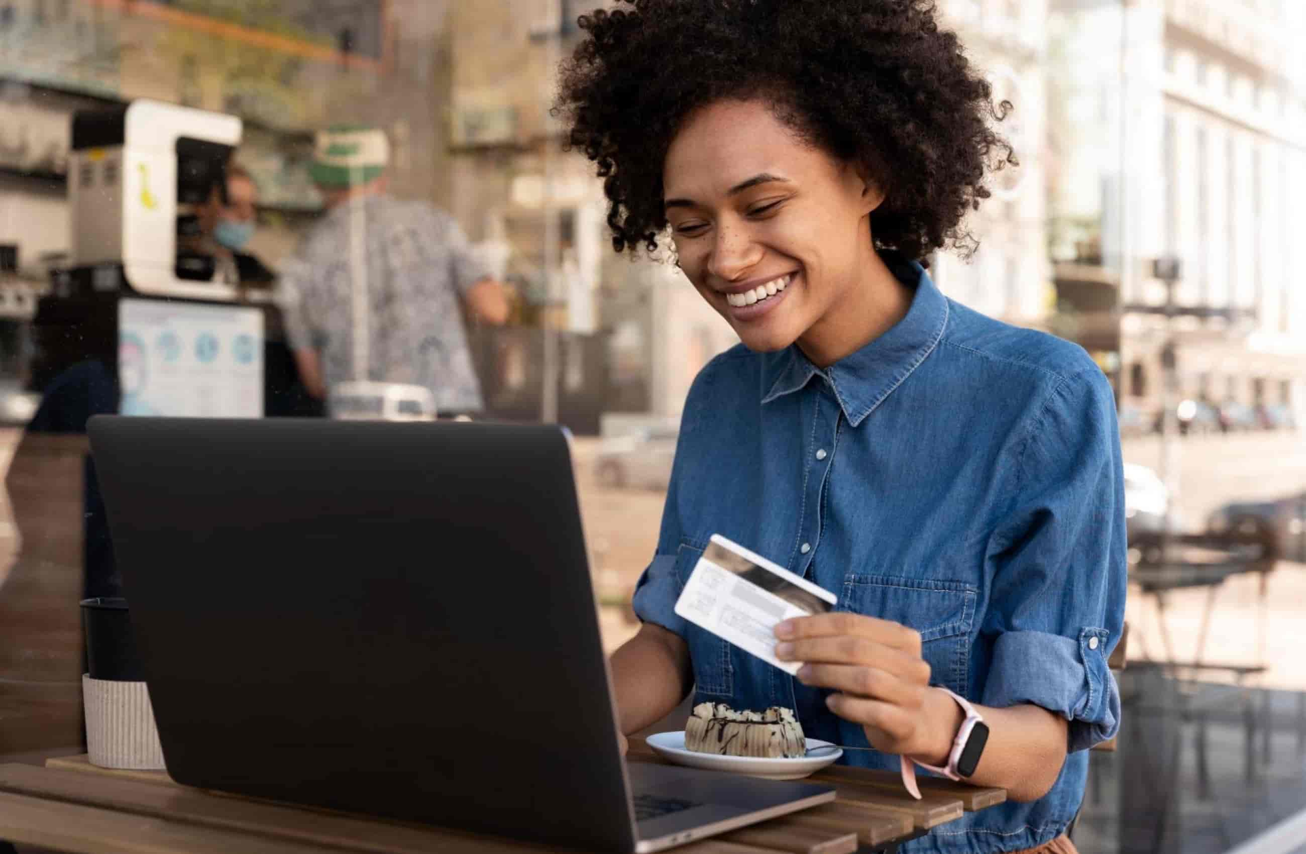 Mulher sentada em um café, vestindo uma camisa jeans. Enquanto segura um cartão com a mão esquerda, usa um notebook para fazer compras online.